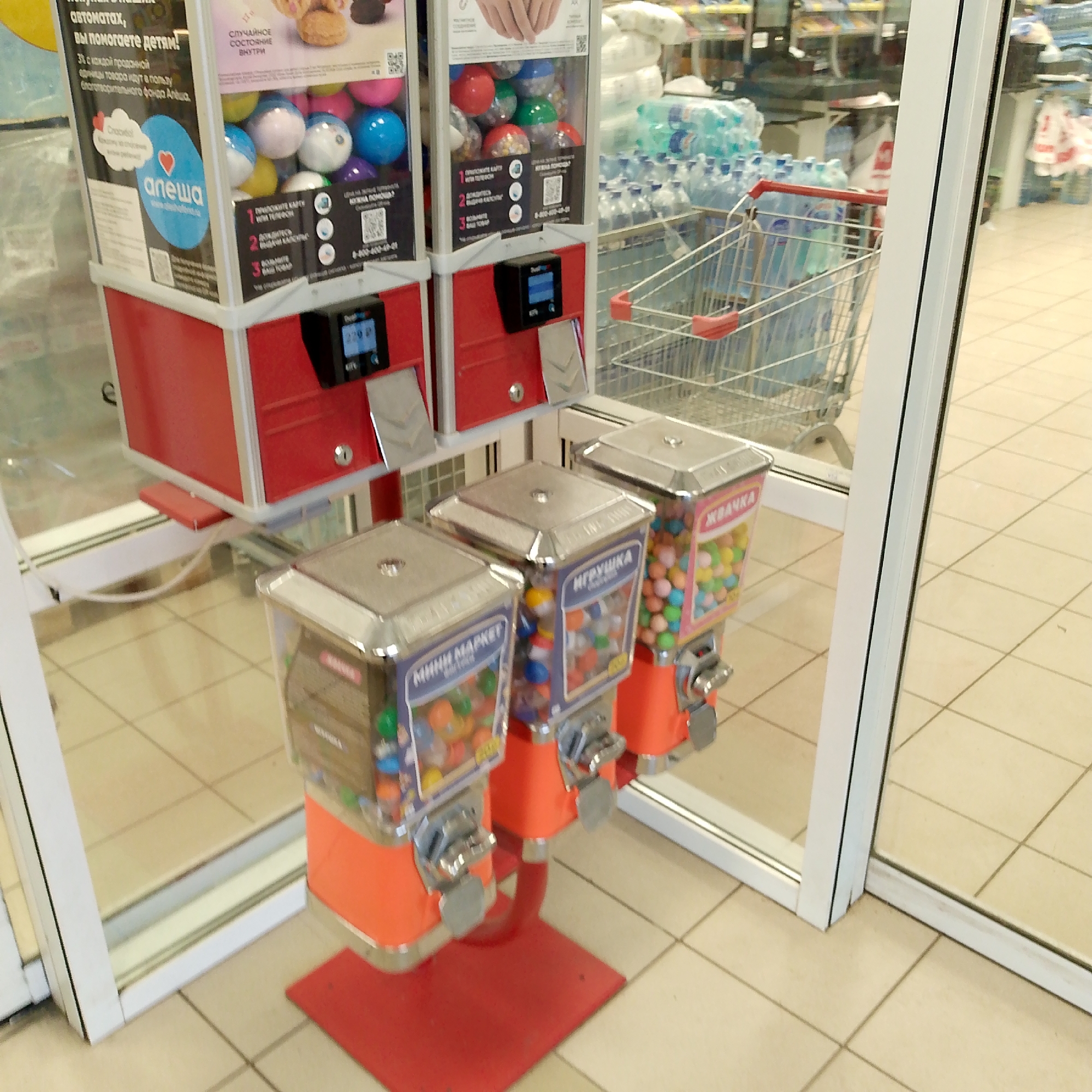 In the city of Nikologory, Russia, a photograph captures a row of gumball machines located inside what appears to be a commercial establishment. The scene is dominated by the bright red and chrome exteriors of the machines, with clear plastic containers filled with colorful gumballs. Labels on the machines indicate their contents: "МИНИ MARKET" (Mini Market), "ИГРУШКА" (Toy), and "ЖВАЧКА" (Chewing Gum). The machines are positioned against a backdrop of tiled flooring and what seems to be a storefront or entrance area with glass panels. In the background, shelves stocked with products and a shopping cart are visible, suggesting a retail environment. The lighting is bright, likely from overhead store lights, and the overall impression is one of a well-lit, clean commercial space. No people are present in the frame.