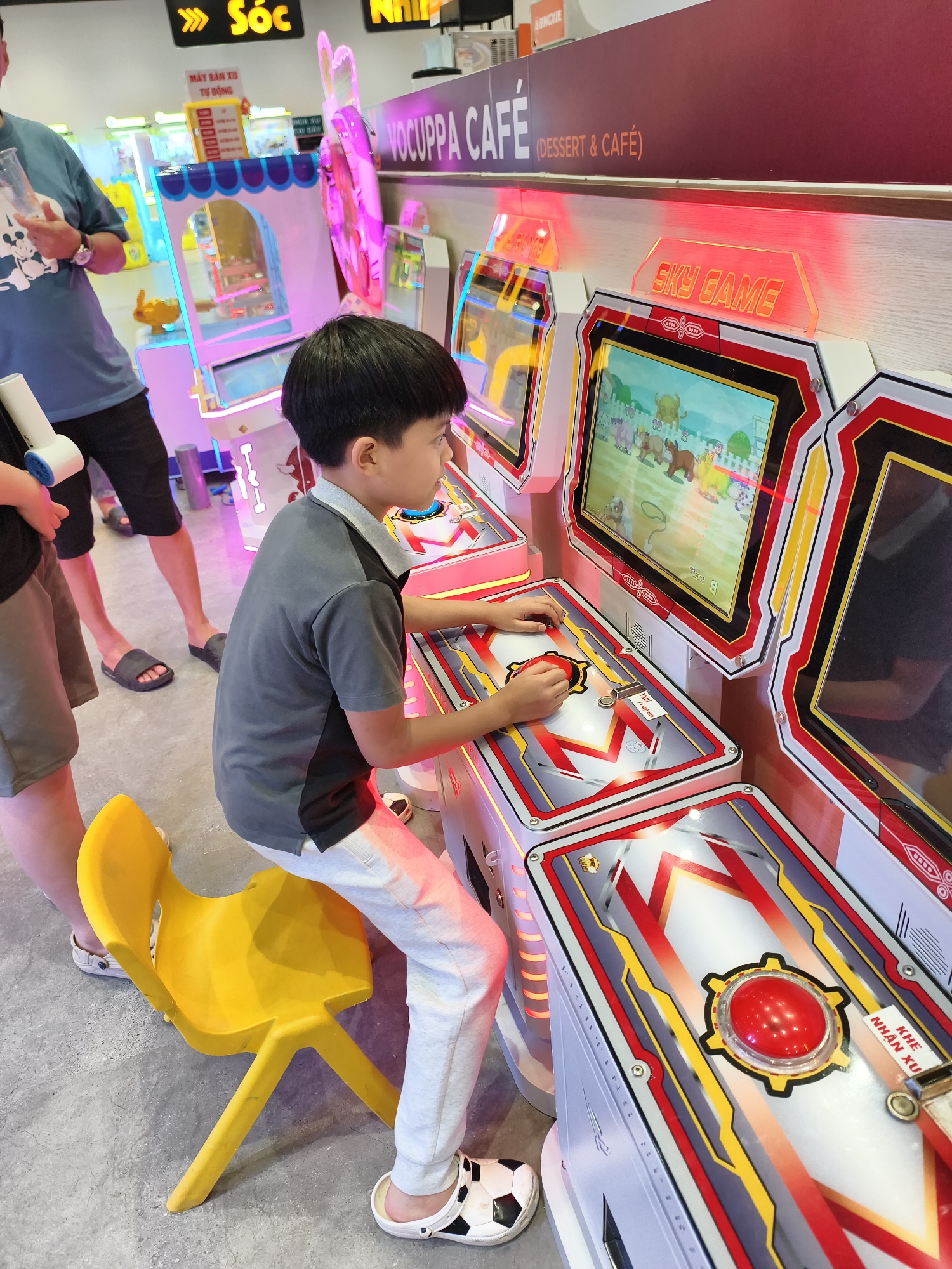 A boy is playing an arcade game called "Sky Game" inside a Vochuppa Cafe, likely in Unknown, Unknown. He is seated on a yellow plastic chair, wearing a gray polo shirt, light gray pants, and soccer-themed sandals. The arcade game features a screen with cartoon animals. Other people are visible in the background, near another arcade game with a blue and pink frame. The cafe has bright lighting and a modern interior design.