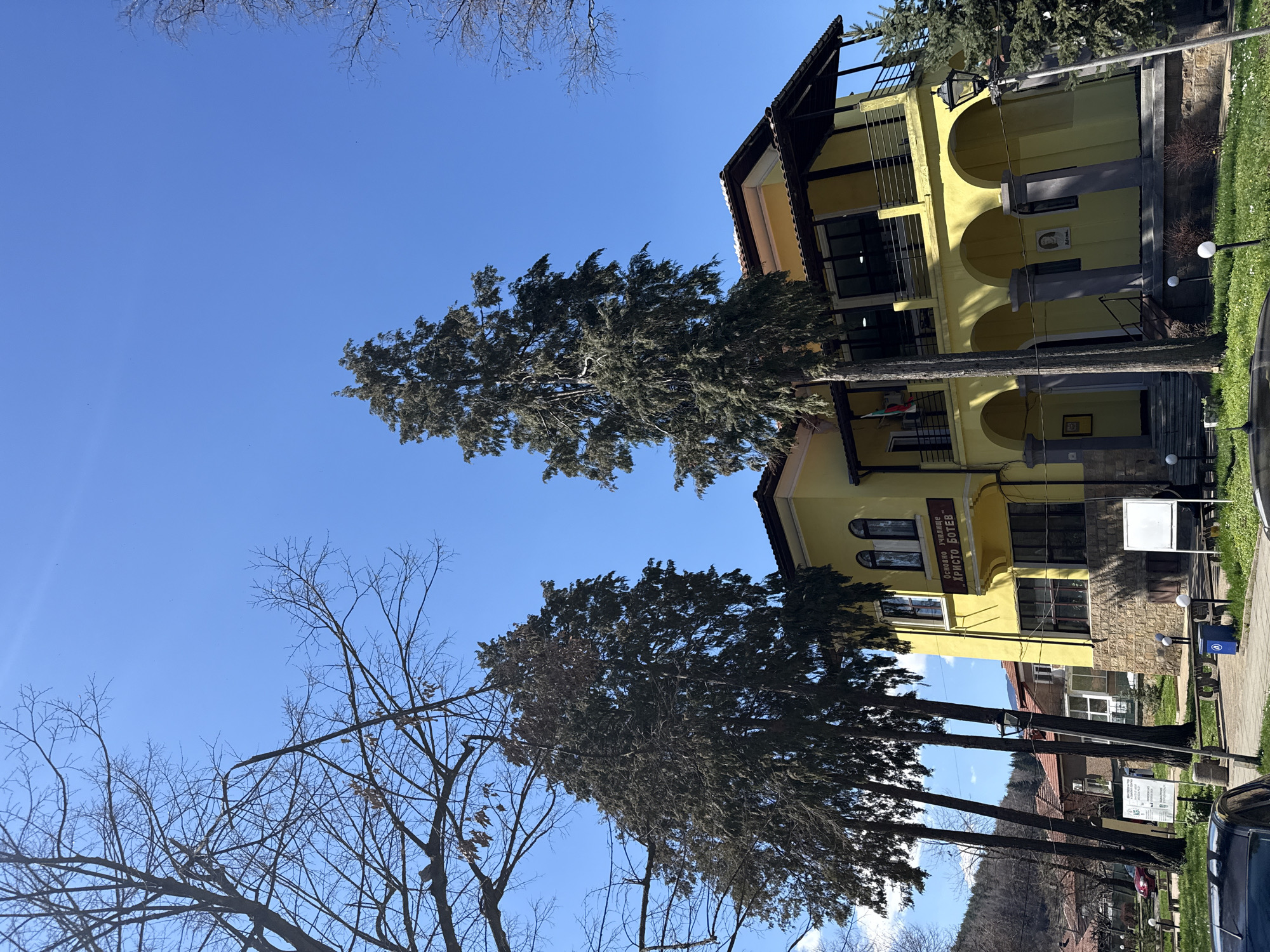 The image shows a yellow building with arched walkways in Etropole, Bulgaria. Trees line the front of the building, with bare branches visible against a clear blue sky. Signage on the building includes the Cyrillic text "ОСНОВНО УЧИЛИЩЕ ХРИСТО БОТЕВ". The location is in the town of Etropole, in the country of Bulgaria.