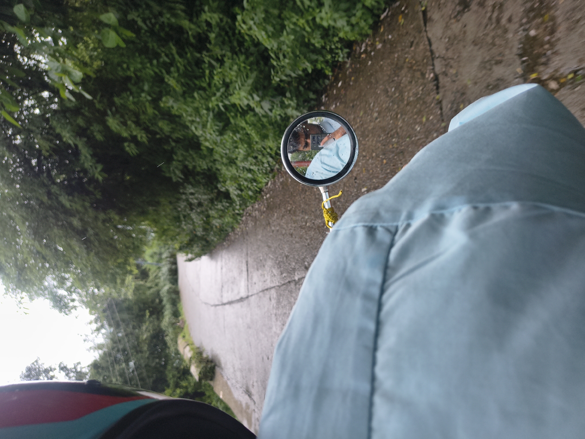 An over-the-shoulder perspective shot shows a person riding a motorcycle on a wet, paved road in Dharamshala, India. The rider is wearing a light blue shirt and a helmet partially visible on the left side of the frame. The road curves to the left, flanked by dense green foliage and trees on both sides. In the circular rearview mirror mounted on the motorcycle's handlebar, a man is reflected, holding a smartphone and taking a selfie. The sky is overcast, and the overall atmosphere suggests a humid, possibly rainy day in a lush, natural setting.