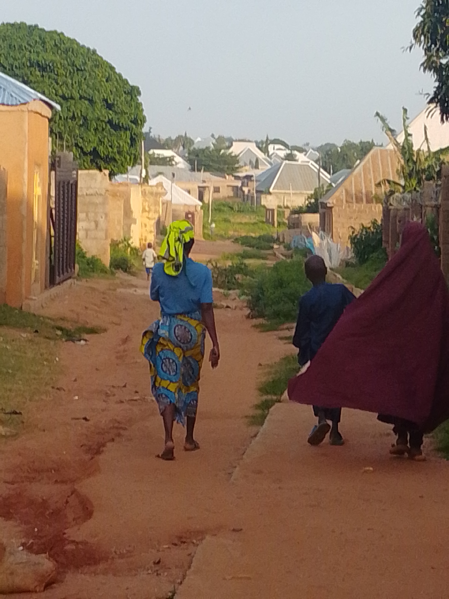 A dirt road runs through a residential area in Gombe, Nigeria, with buildings constructed from tan bricks and concrete. A woman in a blue shirt and a brightly patterned skirt walks away from the camera, her head covered with a yellow and green headscarf. Behind her, a person in a dark blue outfit and a large, deep red cloth walks, partially obscuring them. Further down the road, another person in a dark blue outfit walks away, and in the distance, a child in a white shirt runs. Lush green trees and vegetation line the sides of the road, and more buildings with various roof styles are visible in the background, suggesting a densely populated urban environment. The sky is clear and pale, indicating daytime.