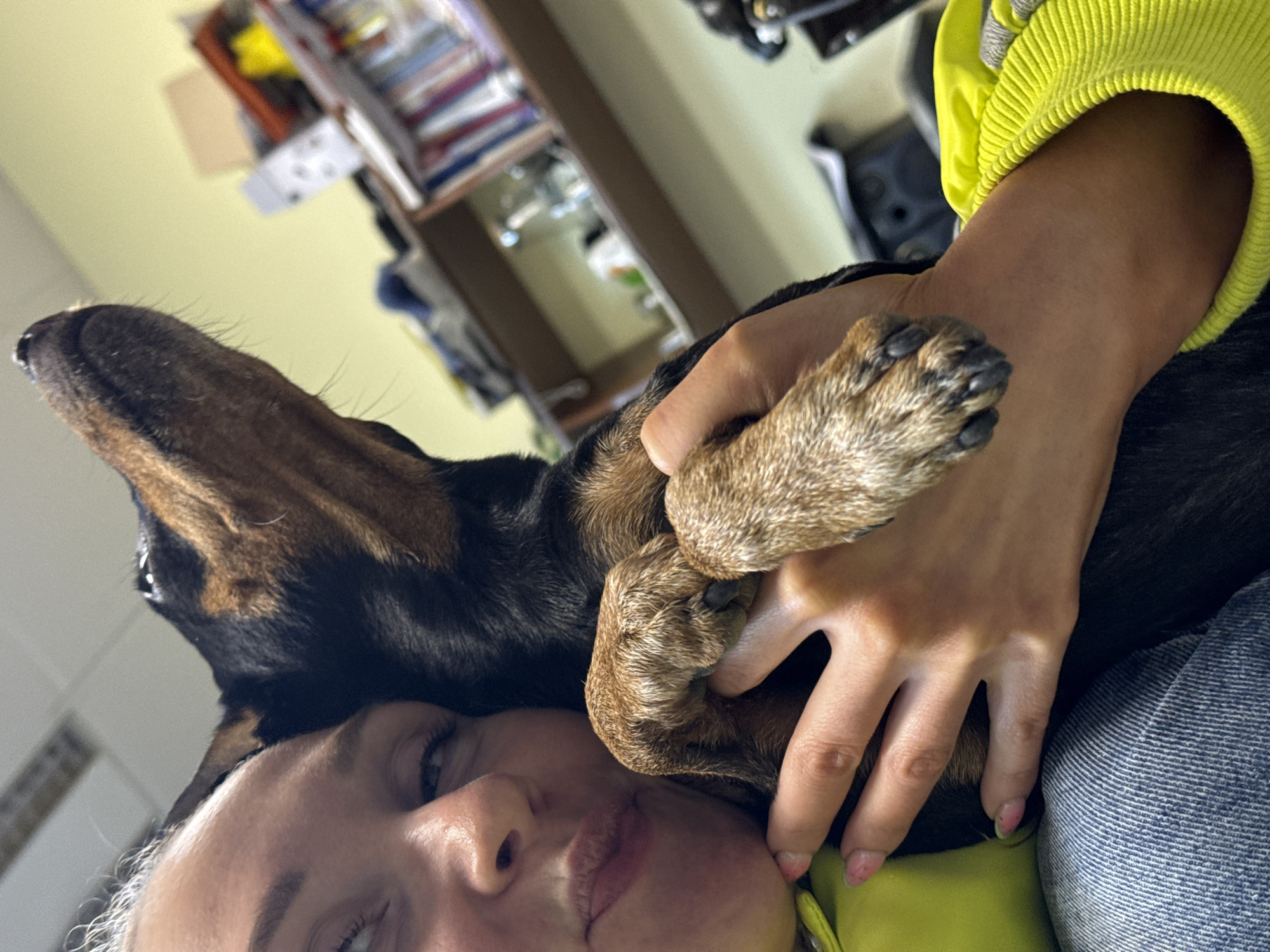 A close-up, tilted shot features a woman with light skin and hair. She is partially visible, with her face and parts of her body in the frame. Her eyes are open, and she is looking slightly to the side. Resting on her face and body is a dog with black and brown fur. The dog's paw is positioned near the woman's face, with her hand gently placed over the paw. The woman is wearing a bright yellow top and jeans. The background is out of focus, showing a room interior with shelves and various objects. There is no definitive information on the location being in the city of Perm, Russia.