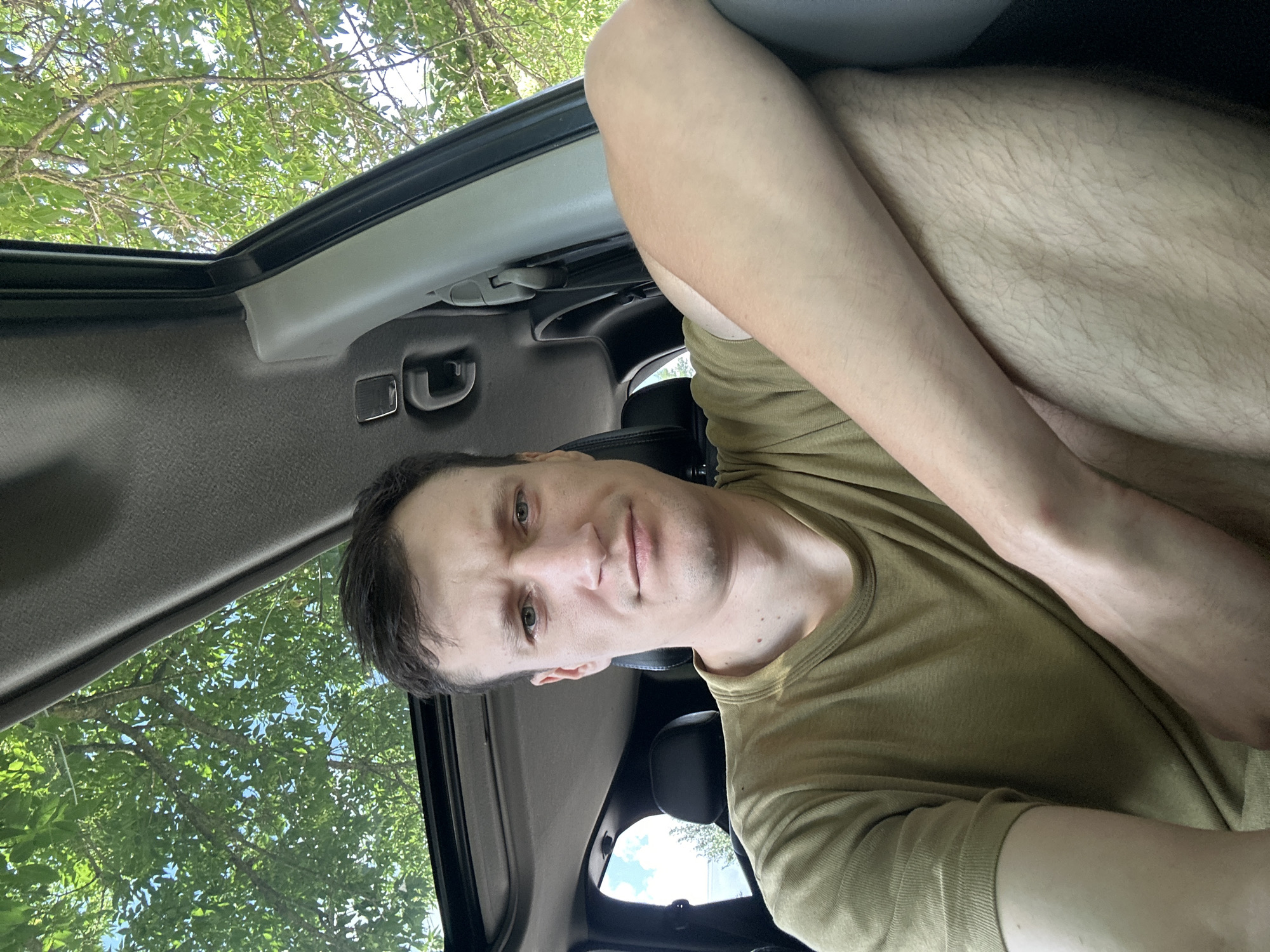 A man is sitting in the passenger seat of a car, looking at the camera. He is wearing a green t-shirt and his right arm is resting on his left leg. The car has a sunroof, and through it, green trees and blue sky are visible. The interior of the car, including the ceiling and seats, is also visible. The man appears to be in a rural or suburban area, possibly in Makiv, Ukraine, given the surrounding vegetation.