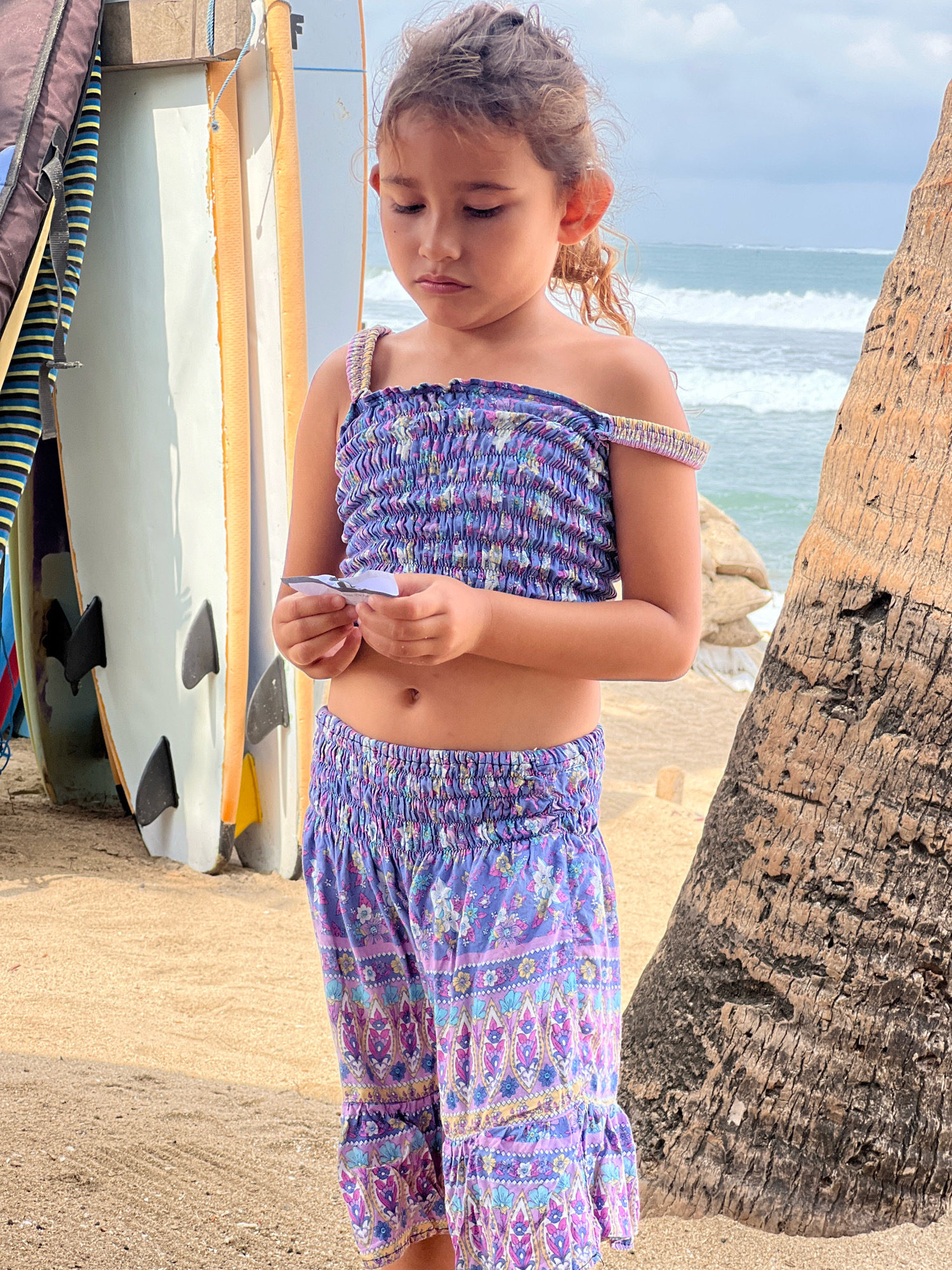 A young girl with curly brown hair is standing on a sandy beach in Kuta, Indonesia, wearing a purple floral print smocked crop top and matching ruffled shorts. She is holding a small piece of paper in her hands, looking down at it with a pensive expression. Behind her, surfboards are leaning against a vertical object, and the ocean with waves is visible in the distance. A palm tree trunk is to her right. The lighting suggests it is daytime, with sunlight casting shadows.