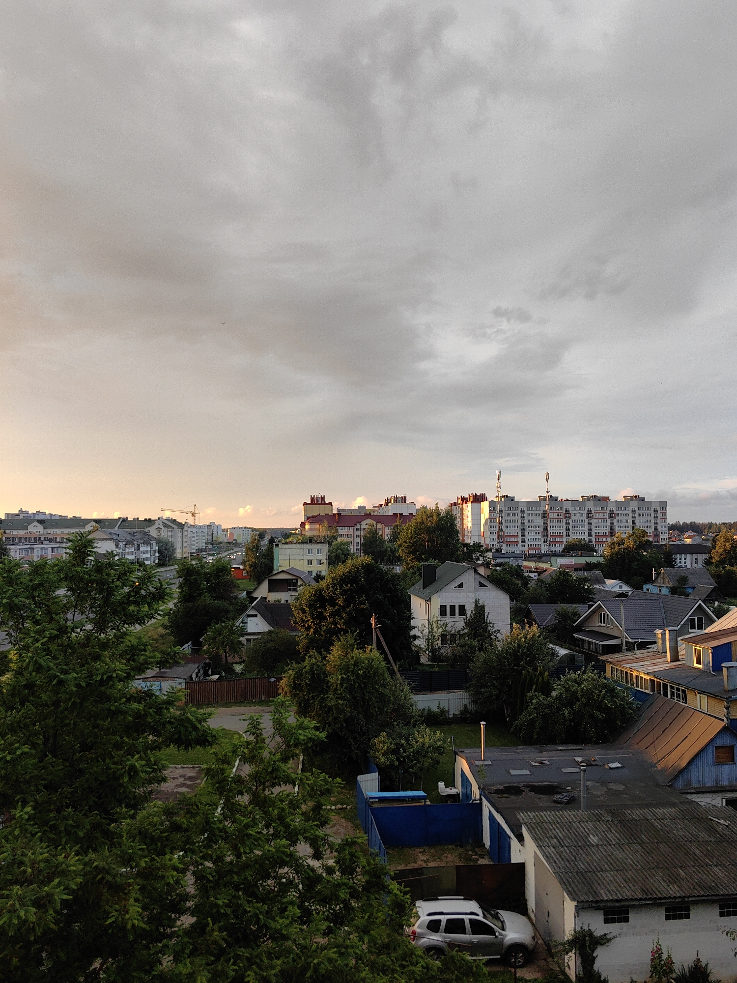 The image depicts a panoramic view of Fanipal, Belarus, during what appears to be late afternoon or early evening. The sky is overcast with a soft, warm light, suggesting either sunrise or sunset. In the foreground, lush green trees obscure the lower portion of the scene. As the view recedes, a mix of residential buildings is visible, ranging from smaller houses with pitched roofs to larger, multi-story apartment blocks.
The apartment buildings are modern, characterized by numerous windows and balconies. One prominent building has a distinctive red and white facade. A construction crane is visible in the middle ground, indicating ongoing development. The environment is predominantly urban with scattered trees and green spaces. Several vehicles are parked on the streets and in driveways, including a silver SUV in the lower right quadrant. The overall impression is that of a quiet, residential area within the city of Fanipal, Belarus. There are no visible people in the image.