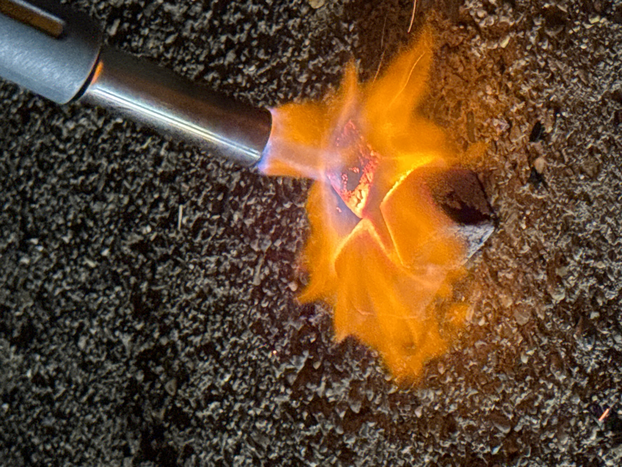 A close-up shot shows a metal blowtorch aimed at a small, dark, possibly square object resting on a rough, pebbly surface. The torch emits a bright orange flame that envelops the object. Small sparks are visible around the flame. The background is out of focus, but appears to be more of the pebbly surface. No people are visible in the image. There is no indication this image was taken in Nizhny Novgorod, Russia.