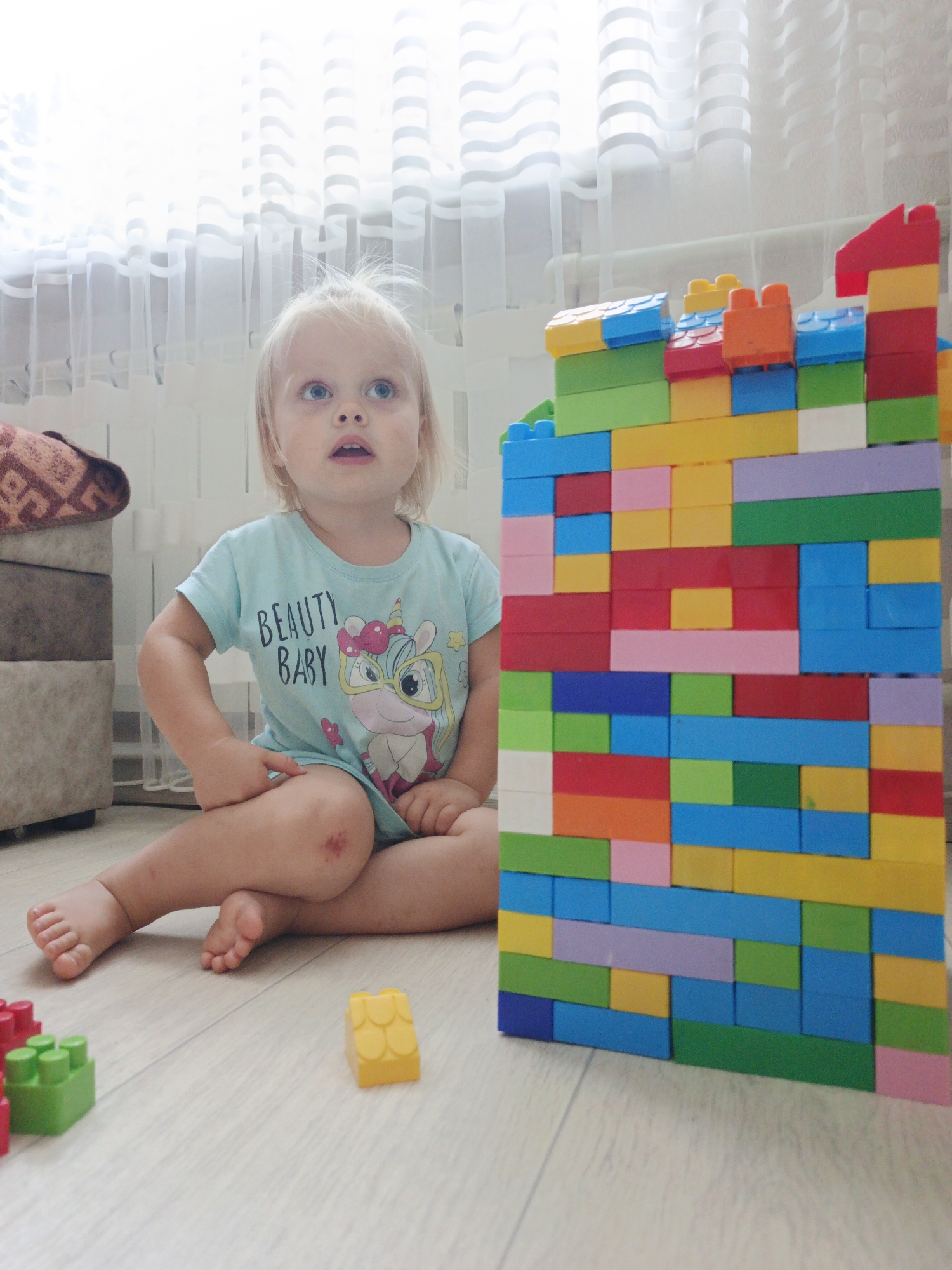 A blond-haired toddler with blue eyes sits on a light wooden floor in front of a wall of colorful building blocks. The toddler is wearing a light blue t-shirt with a unicorn graphic and the words "BEAUTY BABY" printed on it. The toddler's left knee has a red scrape. In the foreground, to the left of the toddler, are a few scattered colorful building blocks. To the right of the toddler and occupying a significant portion of the image is a tall, multi-colored structure made of building blocks. In the background, behind the toddler, is a window with sheer white curtains. The scene appears to be indoors, likely in a home. The image was taken in Zlatoust, Russia.