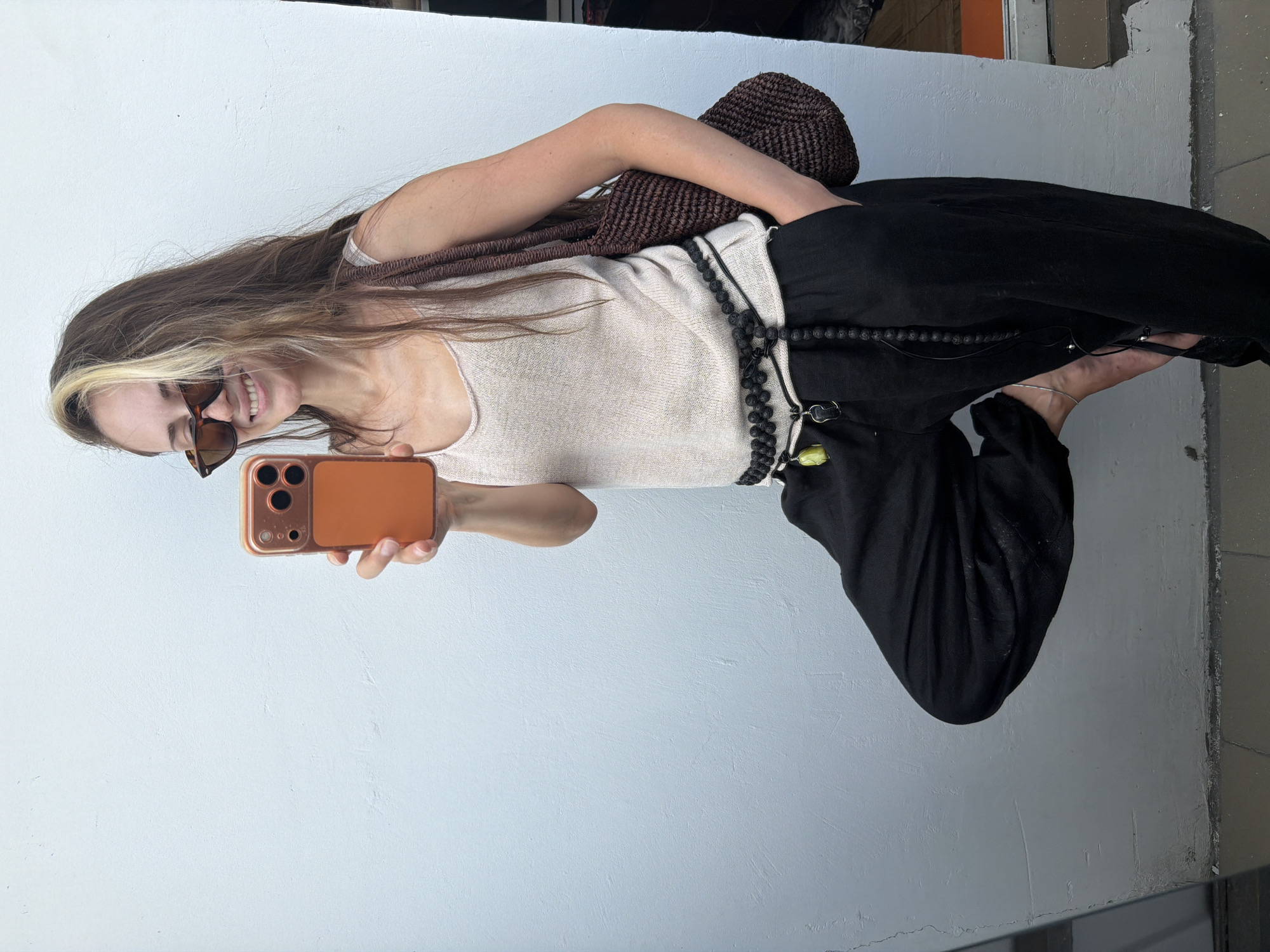 In a softly lit, indoor setting with a light blue textured wall and a tiled floor, a young woman stands with her left leg bent and resting on her right thigh. She holds an orange smartphone in her right hand, seemingly taking a selfie. She wears oversized black pants, a beige sleeveless top, and sunglasses. Around her waist, a black beaded belt is cinched, adorned with a small yellow charm. A brown woven tote bag hangs from her left shoulder. The image does not provide any specific visual cues that would allow for the identification of the location within Khimki, Russia.