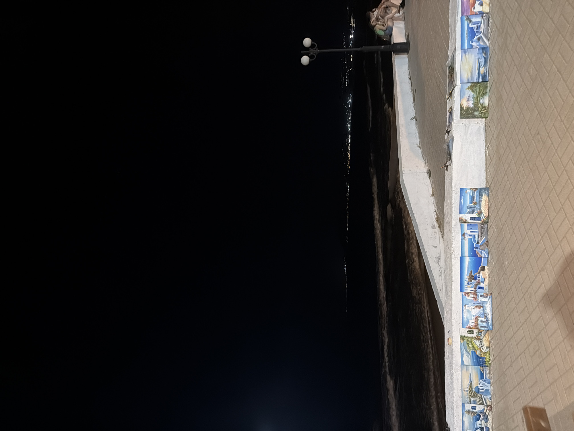 A nighttime view of the coastline in Georgioupoli, Greece, shows a dark sky over a dark sea with faint lights of a distant town visible on the horizon. In the foreground, a white concrete wall runs along the edge of a paved promenade. Along the top of the wall are several small, framed paintings depicting idyllic scenes, mostly of white-washed buildings and blue waters characteristic of Greek islands. A black lamppost with two globes stands to the right of the frame, illuminating a small group of people seated on the wall in the mid-ground. The pavement is made of light-colored interlocking bricks.