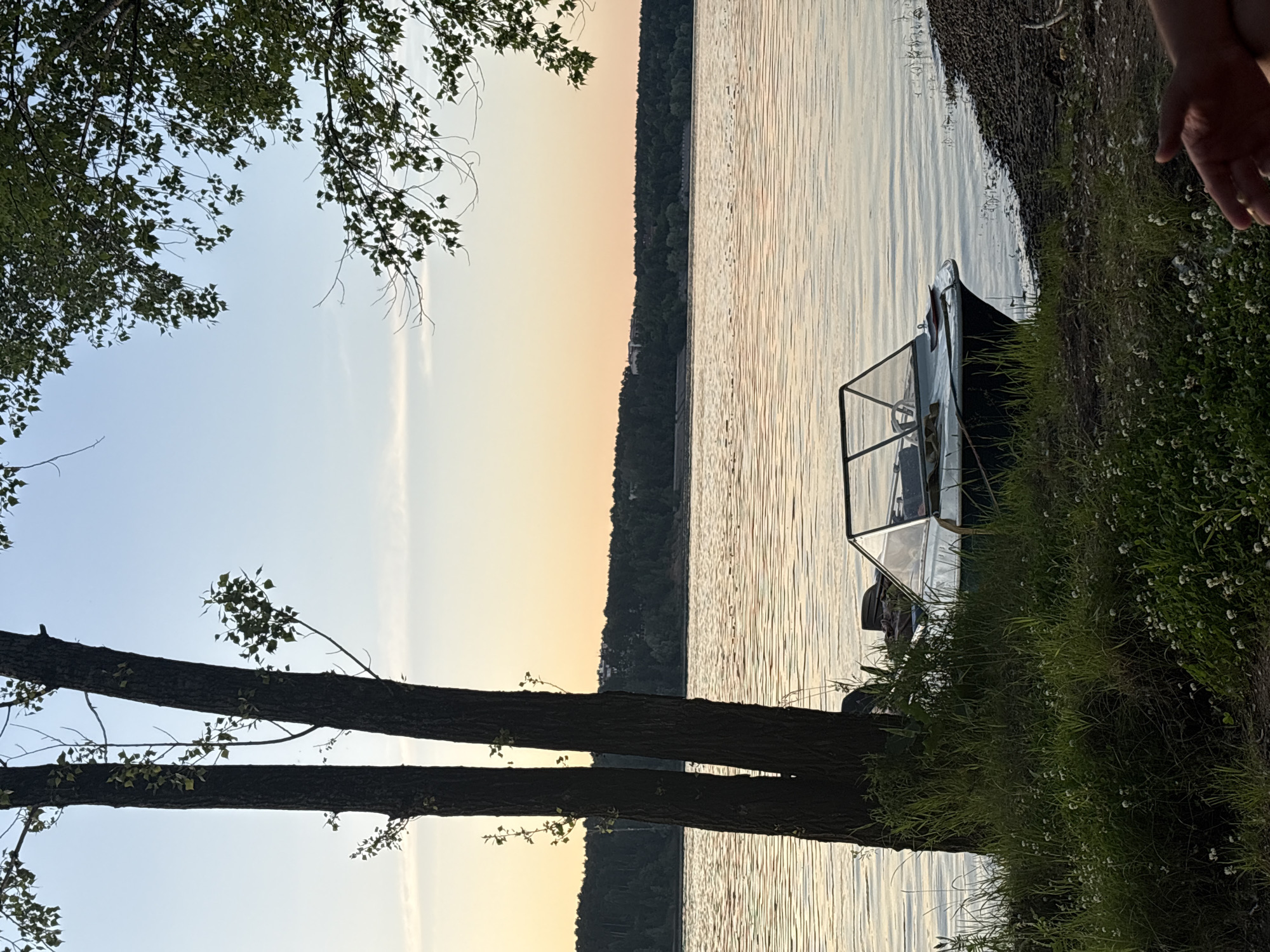 The image depicts a tranquil waterside scene at dusk, likely near the Kama River in the city of Perm, Russia. Two large trees stand prominently on the grassy bank in the foreground, with a small motorboat docked beside them. The boat is dark green with a white windshield and appears empty. The water is calm, reflecting the soft colors of the sunset, with a distant treeline forming the horizon. The sky transitions from a pale blue at the top to a warm orange and yellow hue near the horizon. The overall atmosphere is peaceful and serene, capturing a moment of quiet repose by the water.