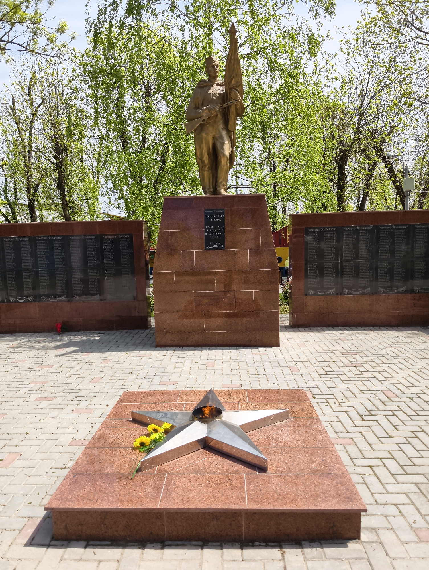 This media file captures a war memorial in Ilskiy, Russia, bathed in bright daylight under a clear sky. The central feature is a bronze statue of a soldier in uniform, holding a flag or banner, standing atop a red granite pedestal. The soldier's posture suggests a solemn and heroic stance.
Flanking the central statue are two tall, rectangular walls made of the same red granite, each featuring several dark plaques inscribed with numerous names, likely of fallen soldiers. Beneath these names, the dates "1941-1945" are visible, indicating that the memorial commemorates those who perished during World War II. A small bunch of red flowers is placed at the base of the left wall.
In front of the central pedestal, on a matching red granite slab, lies a polished silver-colored metal star, designed as an "Eternal Flame," with a small flame burning in its center. A small bouquet of yellow flowers rests beside the star. The ground around the memorial is paved with light-colored bricks laid in a herringbone pattern. Lush green trees, showing fresh spring foliage, frame the background, suggesting a park or public square setting.
The text visible on the central pedestal, in Russian, reads: "ВЕЧНАЯ СЛАВА ГЕРОЯМ" (Eternal Glory to Heroes) followed by "ПАВШИМ В БОРЬБЕ ЗА СВОБОДУ И НЕЗАВИСИМОСТЬ РОДИНЫ" (Who fell in the struggle for the freedom and independence of the Motherland), and again the dates "1941-1945". The scene evokes a sense of remembrance and reverence for the war heroes.