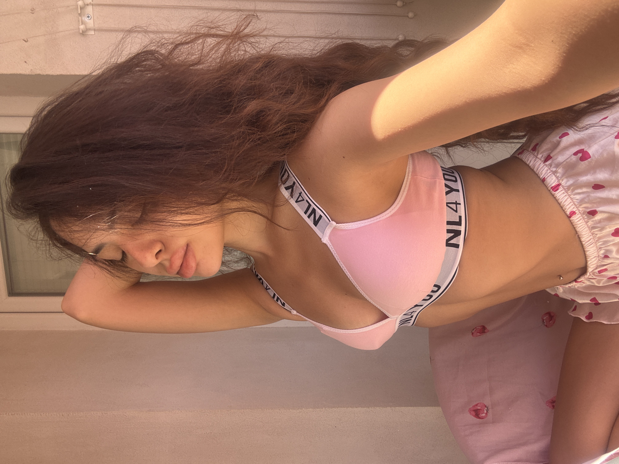 A young woman is sitting in a pink bra and pajama shorts. The bra is light pink with white straps that say "NL4YOU". The shorts are also pink with a heart pattern. She has long, dark, wavy hair and is wearing a silver belly button ring. The woman is indoors, with a window and a wall visible in the background. The scene is lit by sunlight, creating shadows. The location is not specified in the image, and therefore cannot be determined to be in Plovdiv, Bulgaria.