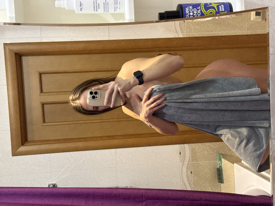 A woman takes a mirror selfie with a phone, partially covered by a gray towel. She wears a black smartwatch. A light brown wooden door is reflected behind her. Bathroom shelves above hold beauty products like "Rag Mary Hydrating Repair Face Tonic" and "Curl Rock and Roll". The bathroom also features off-white tiles with a curved pattern and a purple curtain. A comb rests on a ledge next to the sink.