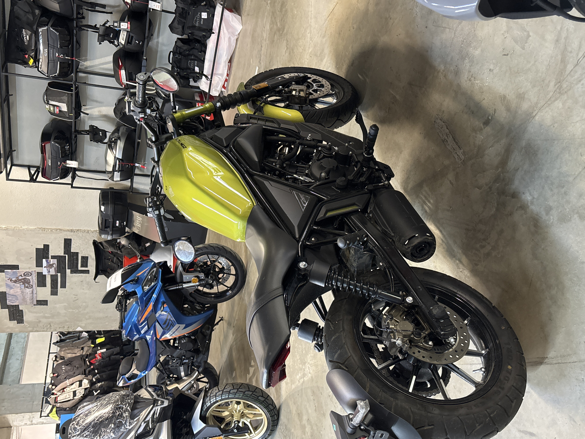 In a brightly lit indoor showroom, likely a motorcycle dealership in Tbilisi, Georgia, a lime green motorcycle is prominently displayed in the foreground, angled slightly to the left. The motorcycle's fuel tank, seat, and rear fender are visible, with the front wheel and handlebars turned to the left. Behind it, a blue and orange sportbike is parked, facing the viewer. To the left of the lime green motorcycle, a silver and black adventure bike is partially visible, with its front wheel and handlebars turned to the left. The background features shelves stocked with various motorcycle luggage, including helmets and saddlebags. A rack of motorcycle riding gear, such as jackets and pants, is also visible on the left side of the image. The floor is a polished concrete surface, with shadows cast by the motorcycles and shelving units. The overall impression is one of a retail environment showcasing a range of motorcycles and accessories.