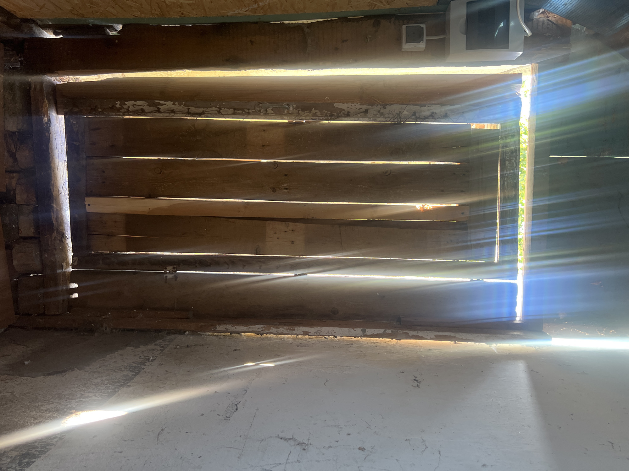 The image shows an interior view of what appears to be an unfinished or derelict building, possibly a shed or a room under construction. The primary subjects are the wooden slats forming a barrier or wall, with visible gaps through which bright sunlight streams in, creating strong beams of light that illuminate the dusty floor and walls. These slats are horizontal and appear aged, with some peeling paint or wood.
The setting is dimly lit, emphasizing the dramatic effect of the sunlight. The floor is concrete and appears rough and dirty, with some lighter patches suggesting previous coverings or repairs. The lighting suggests it is daytime, likely sunny, given the intensity and angle of the sunbeams. The scene evokes a sense of abandonment or a work-in-progress, with a raw, unrefined aesthetic.
Notable details include the texture of the weathered wood, the dust particles visible in the light, and the stark contrast between the bright exterior light and the dark interior. There are some hints of electrical fixtures or conduits on the wall near the top of the frame, suggesting it might be a functional space at some point. No people are visible in the image. The overall mood is one of quiet desolation and stark natural illumination. There is no visible text.