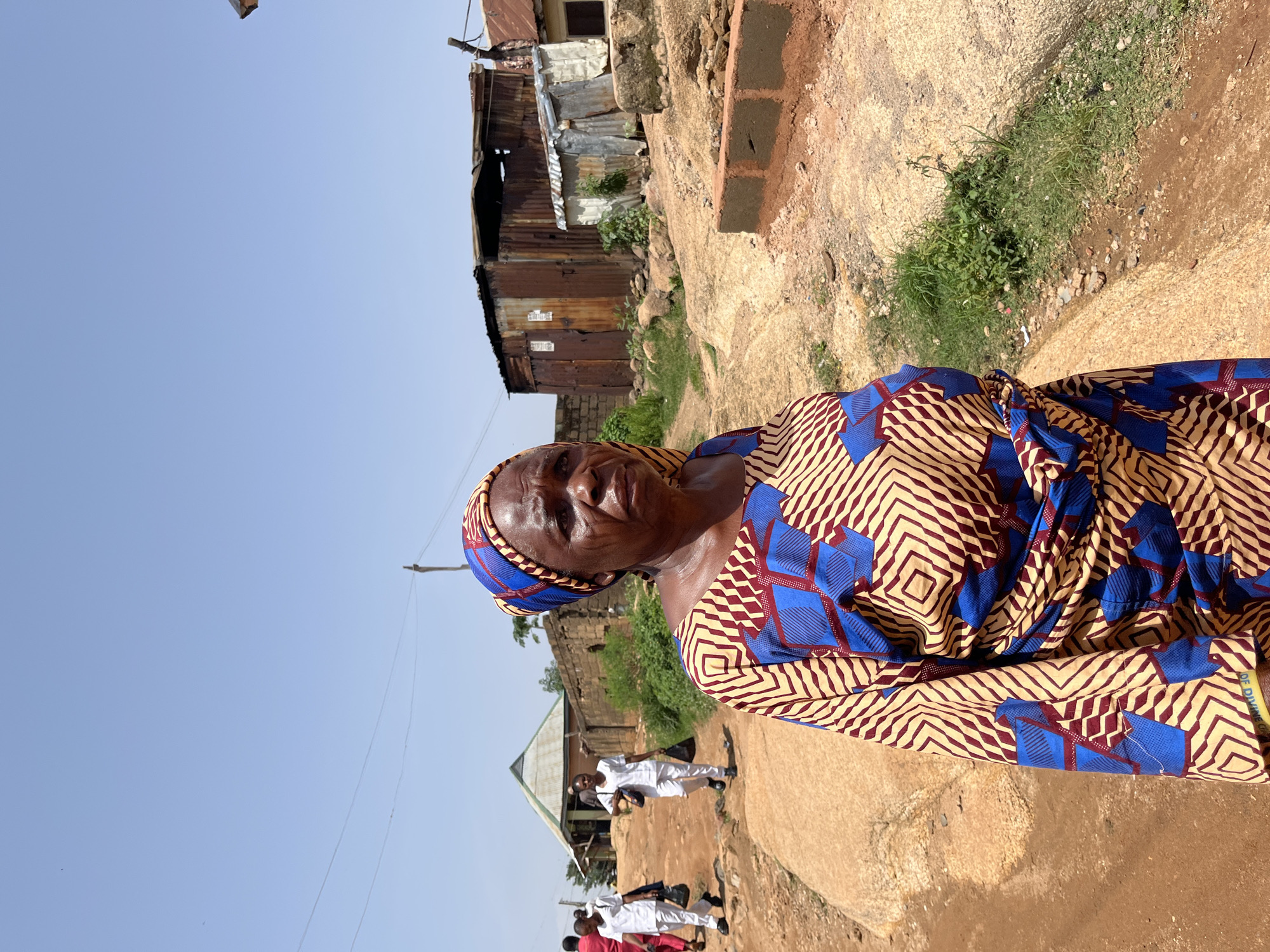 A medium shot captures a woman in the foreground, facing slightly to the right, with her gaze directed forward. She is wearing a headscarf and a vibrantly patterned dress in yellow, blue, and red. Her expression appears somber. The background reveals a dirt road in what appears to be a village or semi-urban area in Jos, Nigeria. Several people in white attire are walking in the distance on the left side of the road, some carrying bags. The environment is dry and dusty, with some scattered vegetation. Several structures, including buildings with corrugated metal roofs and brick walls, are visible. The sky is clear and blue.