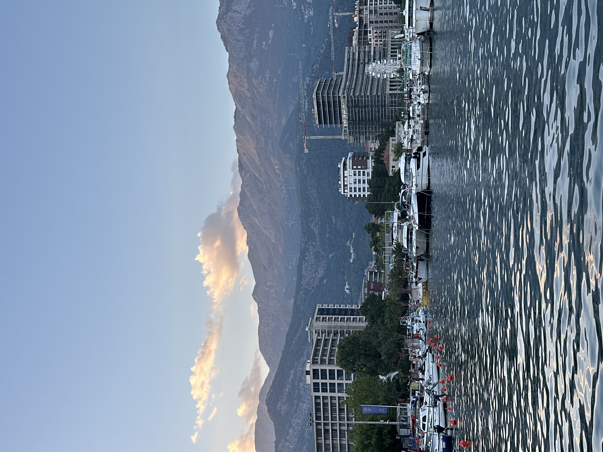 A serene harbor scene unfolds in Budva, Montenegro, as twilight descends. The foreground is dominated by the shimmering surface of the water, reflecting the subdued light and creating a textured, dynamic pattern. A line of orange buoys marks a boundary within the harbor, with numerous boats of varying sizes docked in the midground. These vessels, predominantly white and sleek, bob gently in the calm waters, their masts and superstructures creating a picturesque maritime landscape.
Behind the harbor, the city of Budva emerges, a blend of modern architecture and natural splendor. Tall, contemporary buildings with clean lines and repetitive windows punctuate the skyline, some appearing to be hotels or residential complexes. Construction cranes are visible amongst some of the newer structures, suggesting ongoing development. Further inland, lush green trees and palm trees add a touch of tropical vibrancy to the urban setting.
The backdrop of the scene is a majestic mountain range, its slopes covered in dense, dark greenery. The mountains rise steeply, their rugged peaks softened by the fading light. A few scattered lights are visible on the mountainside, hinting at settlements nestled within the natural landscape. The sky above is a pale blue, transitioning into a warmer hue near the horizon where soft, elongated clouds are tinged with the golden and pink colors of the setting sun. A faint crescent moon is visible in the upper portion of the sky. The overall atmosphere is one of tranquility and picturesque beauty, a harmonious blend of urban life, nautical activity, and natural grandeur.