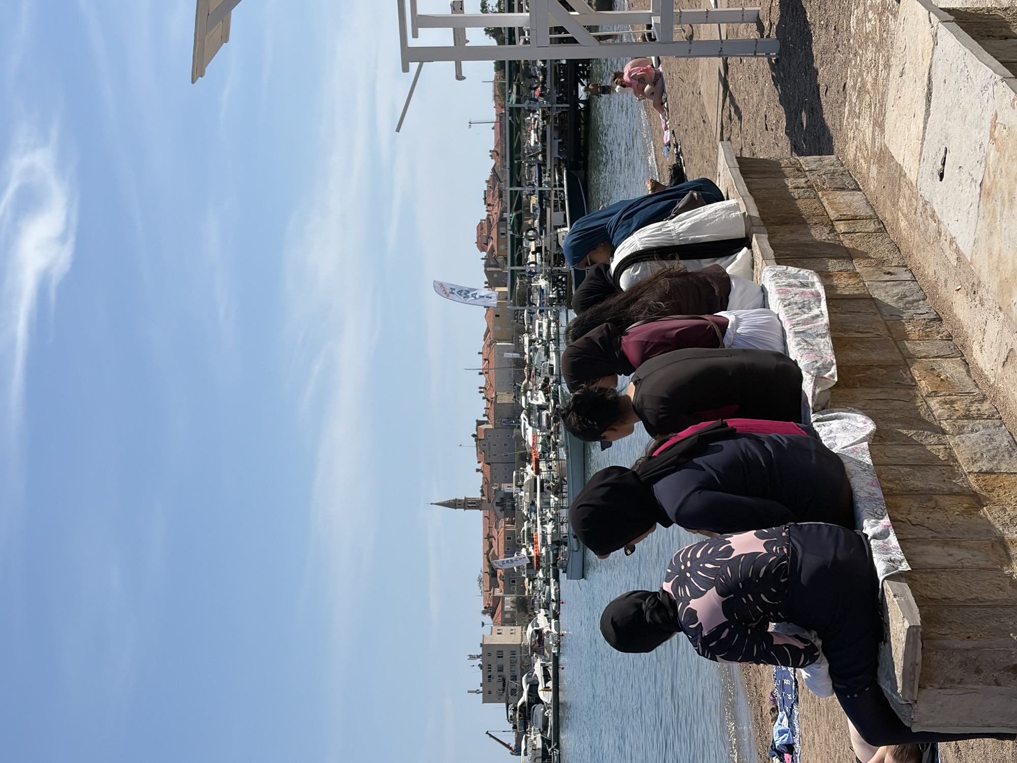 In Budva, Montenegro, a group of six women is seated on a stone ledge facing the Adriatic Sea. The women, from left to right, are wearing full-coverage swimwear, with two wearing headscarves. The woman on the far left is wearing a black full-coverage swimsuit with a pink floral pattern. The next woman is wearing a navy blue full-coverage swimsuit and a black headscarf. The third woman has a burgundy top and a white headscarf. The fourth woman is wearing a dark top and a white headscarf. The fifth woman is wearing a dark top and a blue headscarf. The sixth woman is wearing a dark top and a black headscarf.
Behind them, a marina is filled with numerous boats. In the distance, the city of Budva is visible, with its characteristic red-tiled roofs and a prominent church spire. The sky is clear and blue, with a few wispy clouds. On the beach to the right, a woman and child are sitting on a towel. A white lifeguard stand is also visible on the right side of the image. The overall scene depicts a sunny day at the beach and marina in Budva.