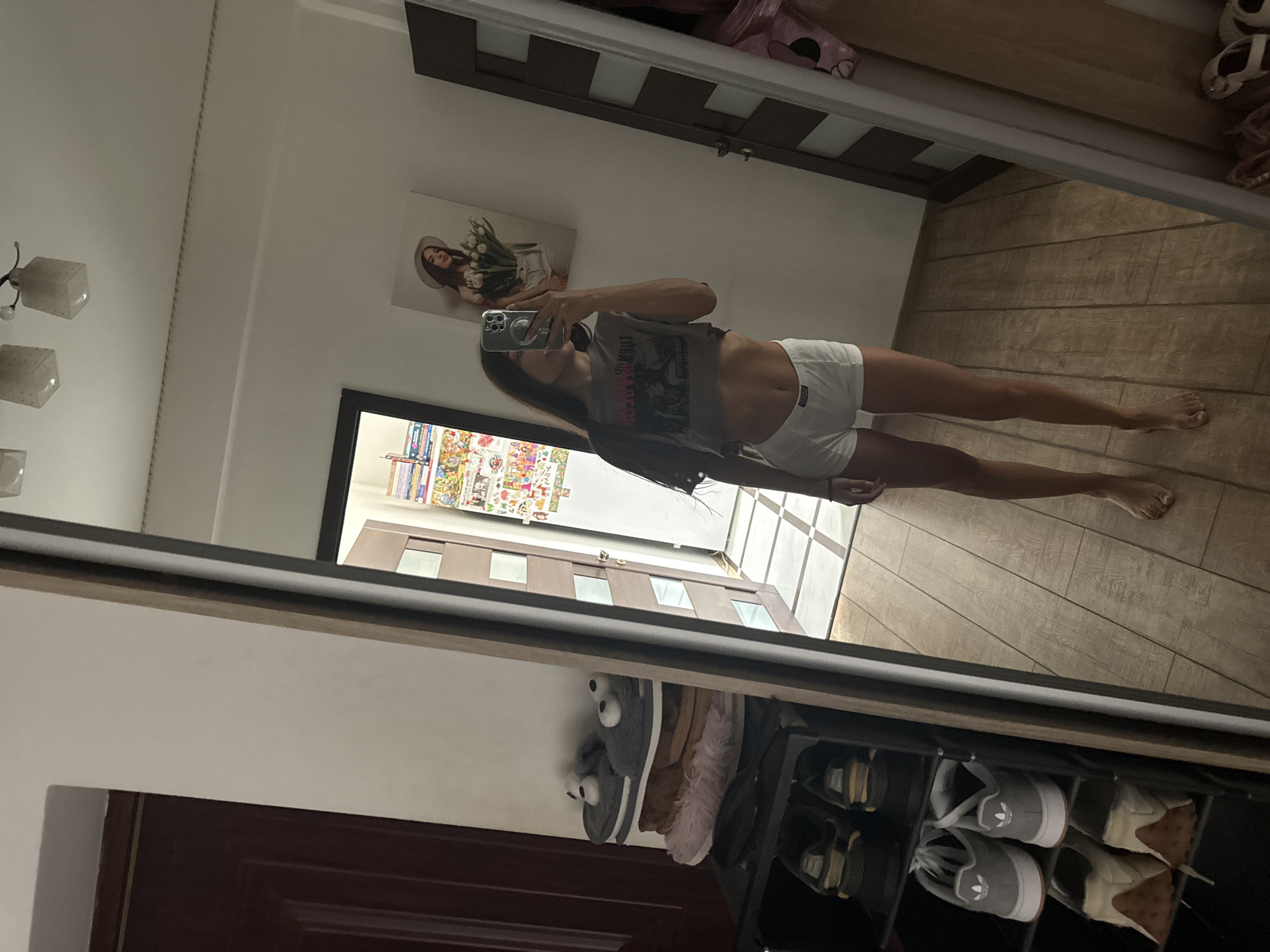 In a room with wood-paneled flooring, a young woman with long dark hair is taking a mirror selfie. She is wearing a gray crop top and white underwear bottoms. In the foreground, a black shoe rack is visible, holding several pairs of shoes, including slippers with bear faces. Behind her, a tall mirror reflects the room, revealing a doorway with glass panes and a wall adorned with a framed picture of a woman holding flowers and numerous colorful stickers. The setting appears to be residential.