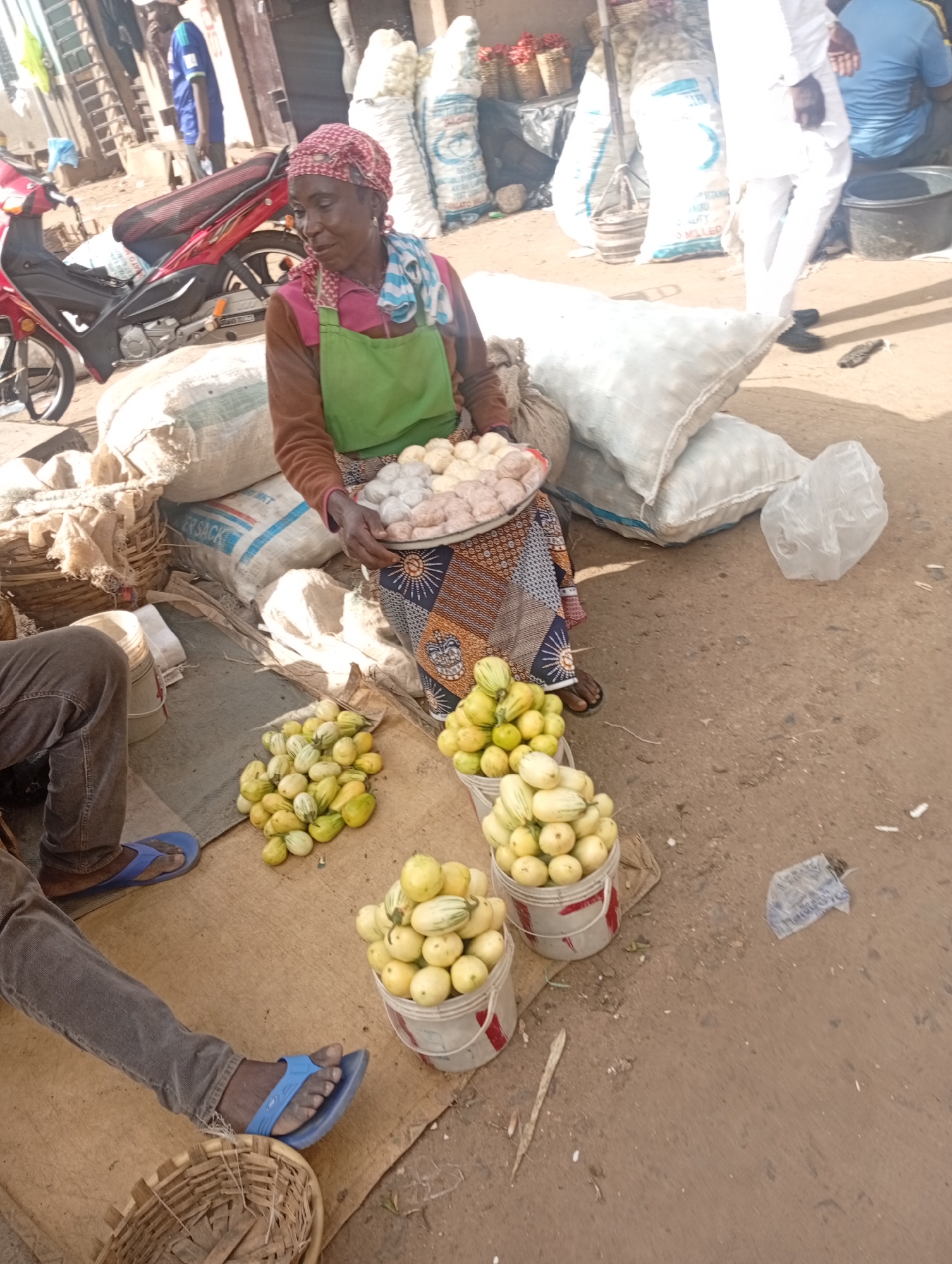 The image captures an outdoor market scene in Rafin Par, Nigeria. A woman wearing a headscarf and an apron stands in the foreground, holding a tray of round, light-colored objects, possibly food items. She is seated next to a large sack of goods and surrounded by other sacks and baskets. In front of her are piles of small, striped gourds or vegetables. To the left, a person wearing jeans and blue flip-flops is partially visible, and a red motorcycle is parked behind the woman. The ground is covered in dirt and debris, typical of a marketplace. In the background, other individuals and stalls suggest a bustling commercial environment. The overall impression is of daily commerce in a Nigerian town.
