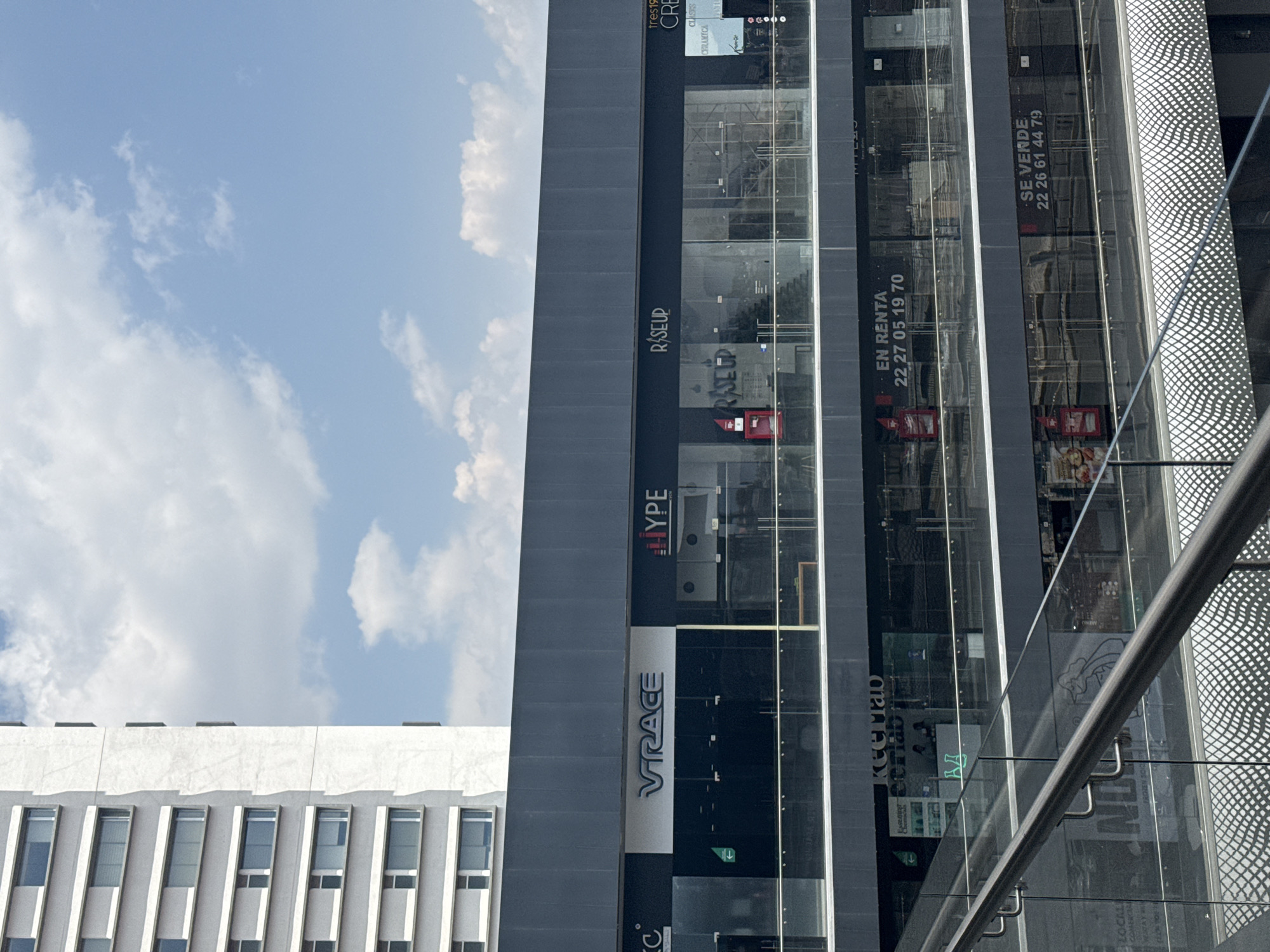 This is an exterior shot of a modern commercial building with a facade featuring glass panels and prominent signage for businesses. The building is multi-story with brands like "VIRACE", "ROSEUP", and "TYPE" clearly visible. A sign indicating "EN RENTA 22 27 05 19 70" (FOR RENT) and "SE VENDE 22 26 61 44 79" (FOR SALE) is present, suggesting real estate opportunities. The building next door is a white building with small rectangular windows. The sky is partly cloudy. The image was taken in San Bernardino Tlaxcalancingo, Mexico.