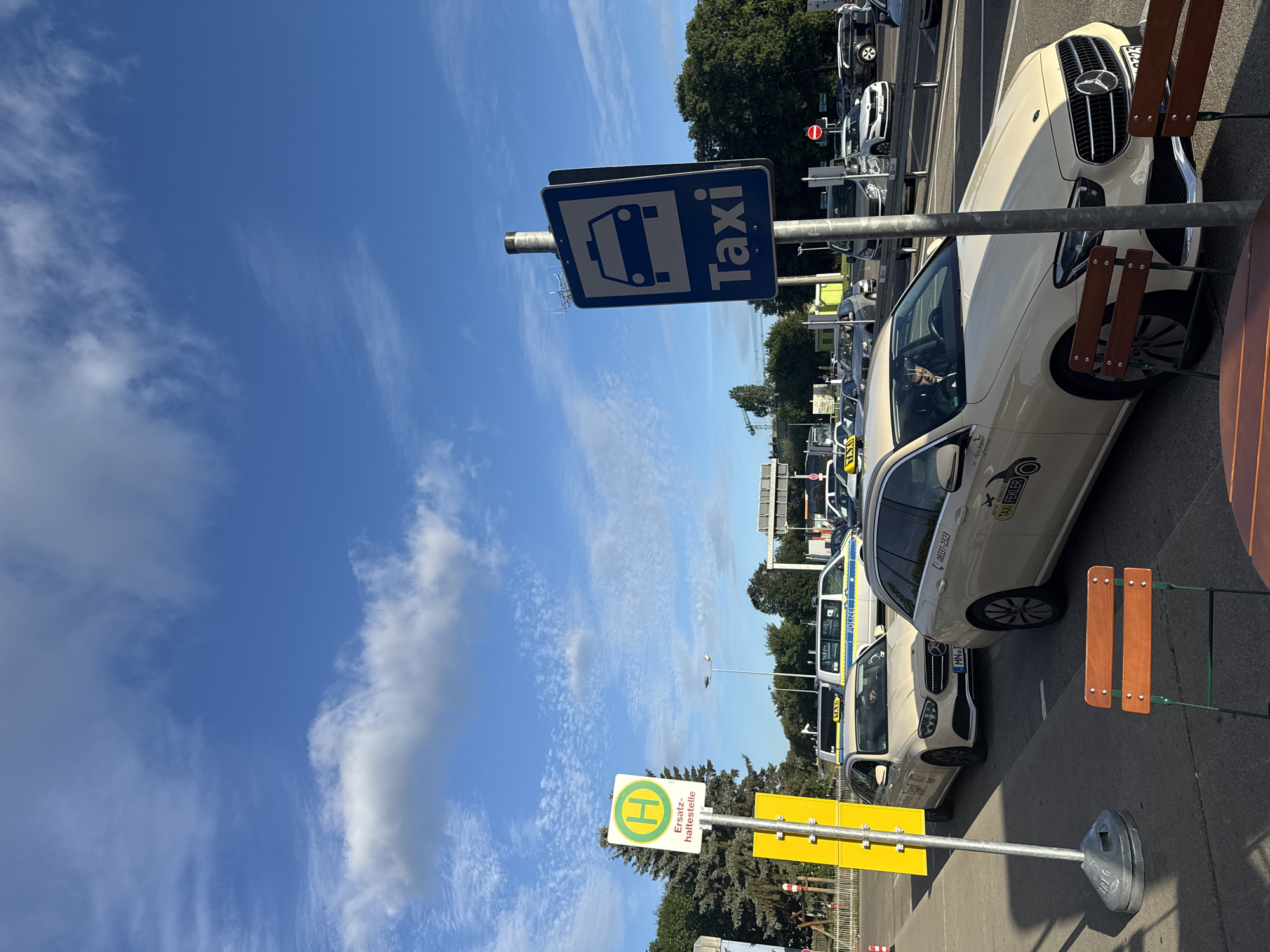 The image captures a bright, sunny day with a clear blue sky dotted with fluffy white clouds, at the Künersberg, Germany, taxi stand. The scene is dominated by several light-colored taxis, likely Mercedes-Benz sedans, lined up in a queue. One taxi in the foreground displays "TAXI FEKLER" on its side and has the phone number "0331-2323" visible. Another taxi further back has a driver behind the wheel. A police van, marked with "POLIZEI," is also present in the lineup.
To the left, a green and white bus stop sign with a yellow "H" symbol and the German text "Ersatz-haltestelle" (temporary stop) stands prominently. Behind the bus stop sign, a dense line of evergreen trees forms a backdrop. In the middle ground, a traffic signal and other vehicles are visible, suggesting a road intersection or a busy transportation hub. In the foreground, two wooden chairs with metal frames are set up, hinting at a waiting area or an outdoor seating space. The overall impression is one of an organized taxi stand in an urban or semi-urban setting on a pleasant day.