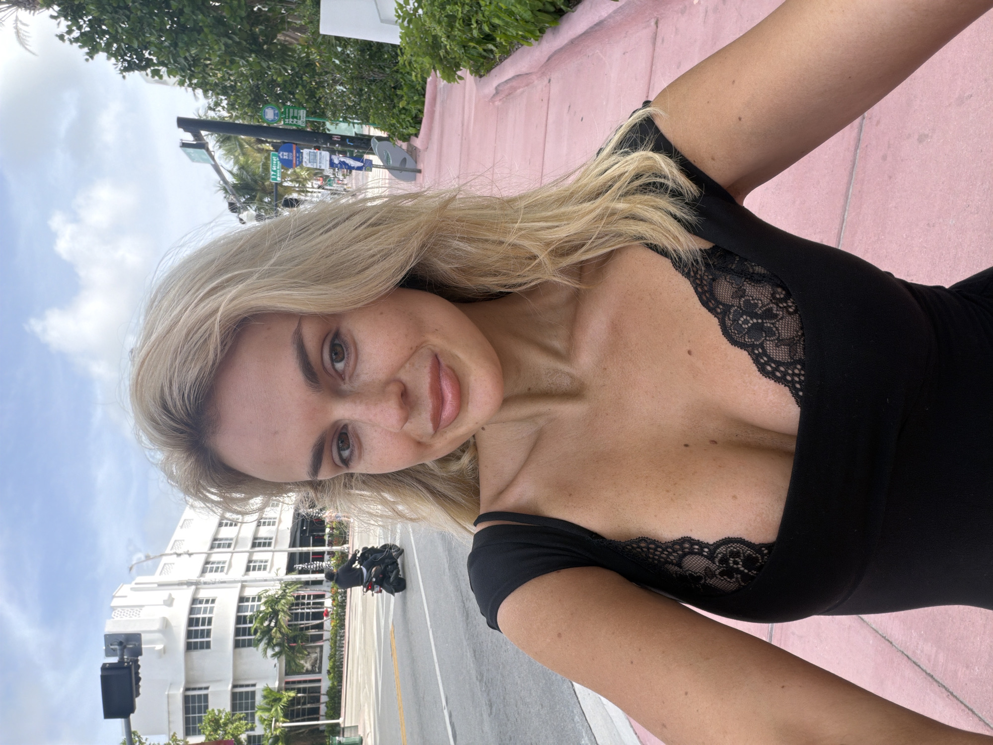 A blonde woman with freckles is taking a selfie on a sidewalk in Miami Beach, United States. She is wearing a black top with lace details around the neckline. The sidewalk is pink, and there is a street and buildings visible in the background. A motorcycle is parked on the street. Street signs indicate "11 Street". Palm trees and other greenery are also present. The sky is partly cloudy.