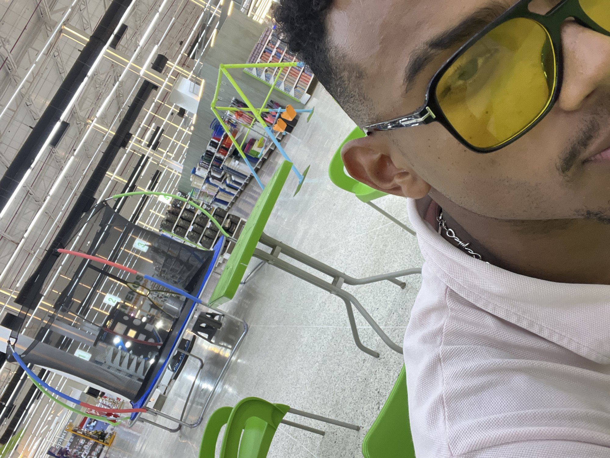 A selfie of a person with yellow sunglasses and a silver chain, wearing a pink shirt, is the focal point. In the background, a trampoline with blue, red, and green accents is visible. There are green tables and chairs. The background suggests a retail environment with visible shelving, likely within the city of Porlamar, Venezuela.