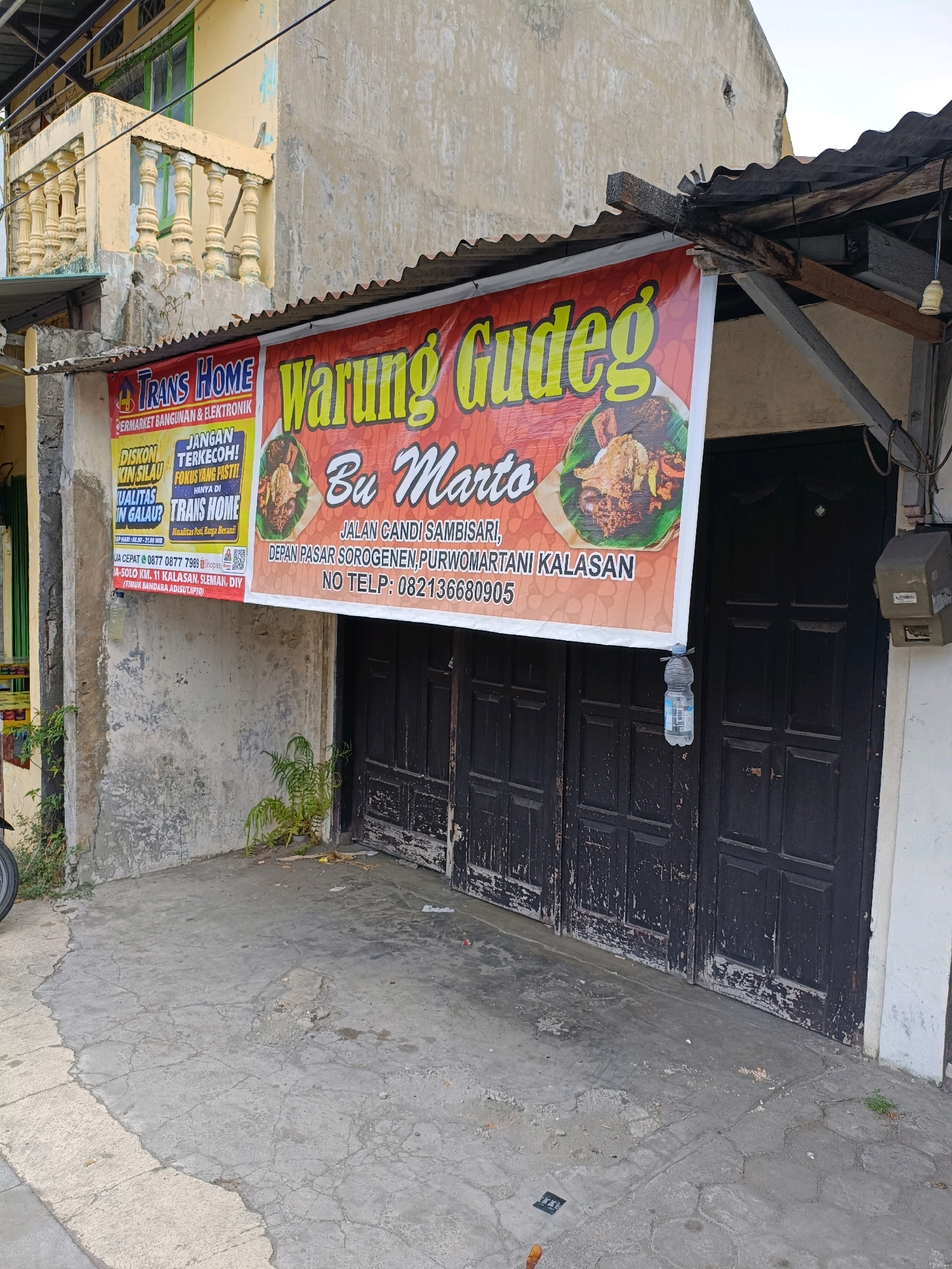 The image shows the exterior of a building with a banner displaying "Warung Gudeg Bu Marto." The banner is red and orange and features a picture of Gudeg, a traditional Javanese dish. Below the restaurant name, the address is listed as "Jalan G Andi Sambisari, Depan Pasar Sorogenen, Purwomartani, Kalasan" and a phone number "082136680905." To the left of the Gudeg banner is another banner for "TRANS HOME," a store selling building materials and electronics. This banner is yellow and red and advertises discounts. The building itself appears weathered, with a textured concrete wall and a corrugated metal roof. Two dark-colored wooden doors are visible, leading into the establishment. A plastic water bottle is hanging from a wire near the doors, possibly to deter flies. Small plants are growing in the cracked concrete area in front of the building. In the background, a yellow building with a balcony and green-framed windows is partially visible. The scene is outdoors, suggesting it is a street-level view of a commercial establishment in Purwomartani, Indonesia.