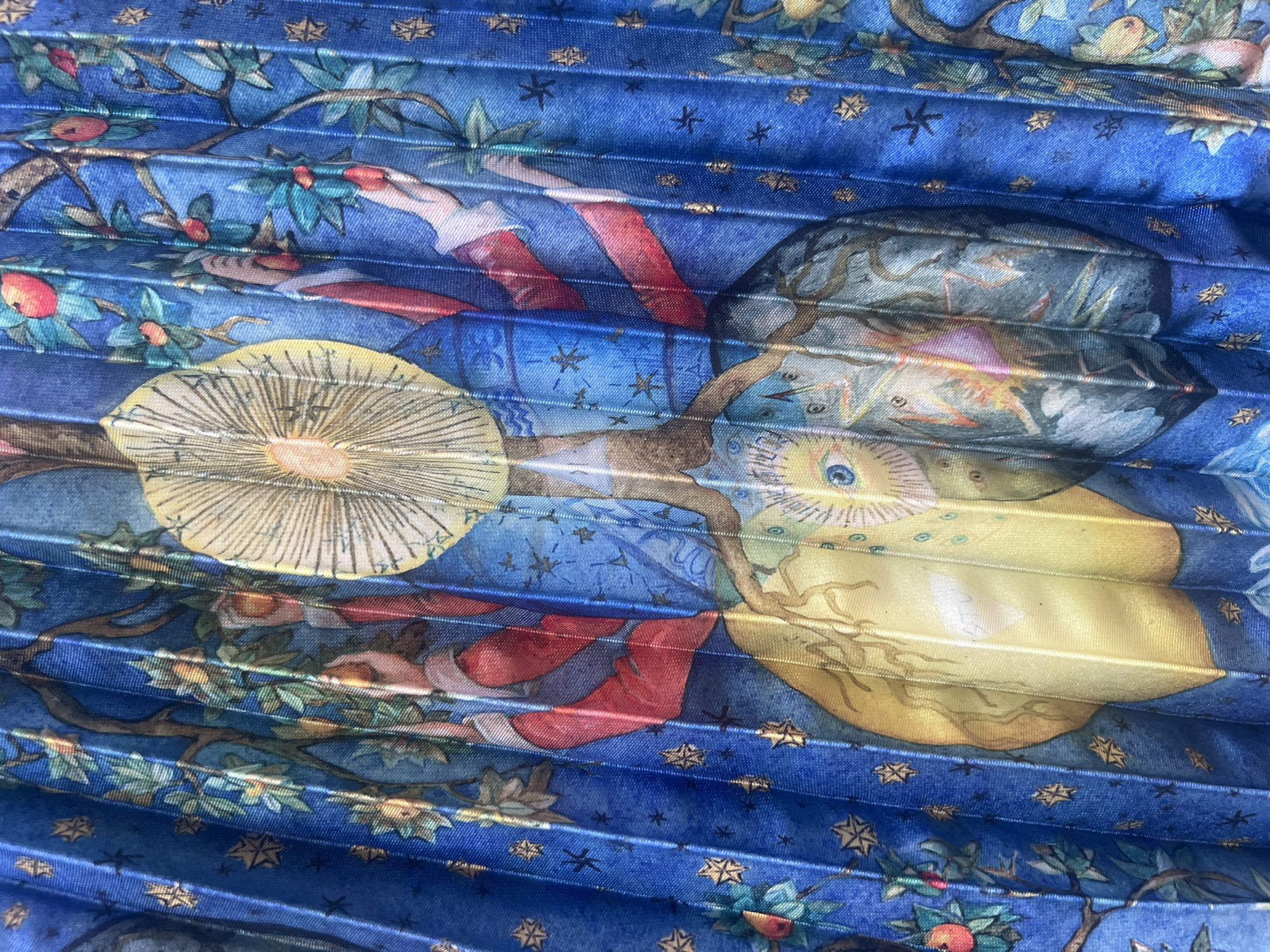 This image is a close-up of a pleated fan with a blue background depicting a fantastical scene. The fan is adorned with a watercolor-style illustration featuring astrological and nature motifs. There are stylized trees with red fruits, celestial bodies like a large yellow sun with rays and a faceted eye, and what appear to be figures in red and white garments reaching for the fruit. Stars and other decorative elements are scattered across the blue expanse. The pleats of the fan create a textured, layered effect, and the metallic sheen of the fabric is visible in the highlights. The image does not contain any elements that would allow for its location to be identified in the city of Mollet Del Vallès, Spain.