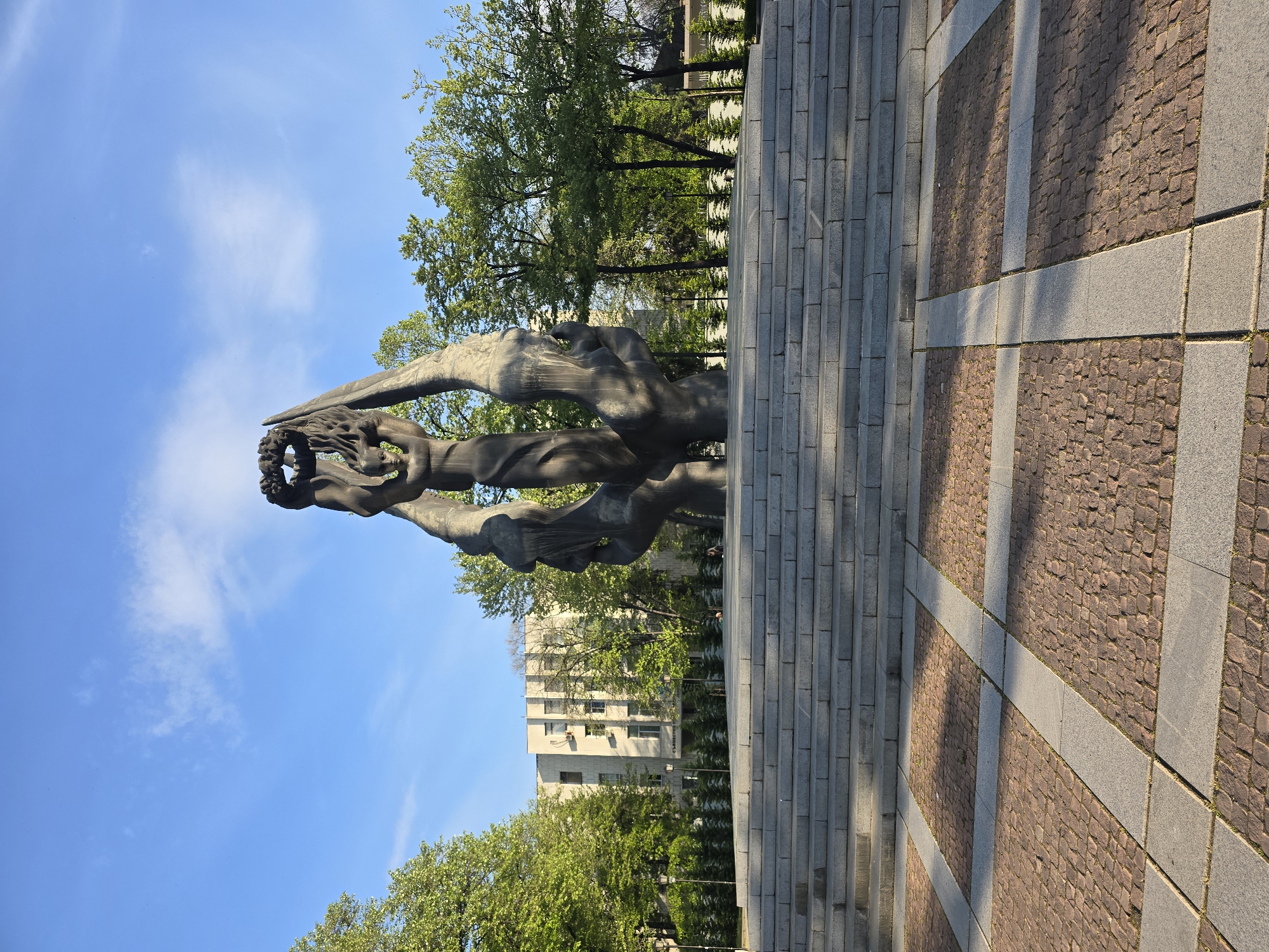 Here is a detailed description of the image:
The image shows a bronze sculpture of two figures with outstretched arms, holding a wreath aloft. The sculpture stands on a gray stone platform or steps, which leads onto a patterned brick surface with gray stone inlays. In the background, there are trees and a tall building. The sky is blue with some scattered clouds. The sculpture is likely a monument or memorial, and the setting appears to be an urban park or square. The location is Unknown, Unknown.