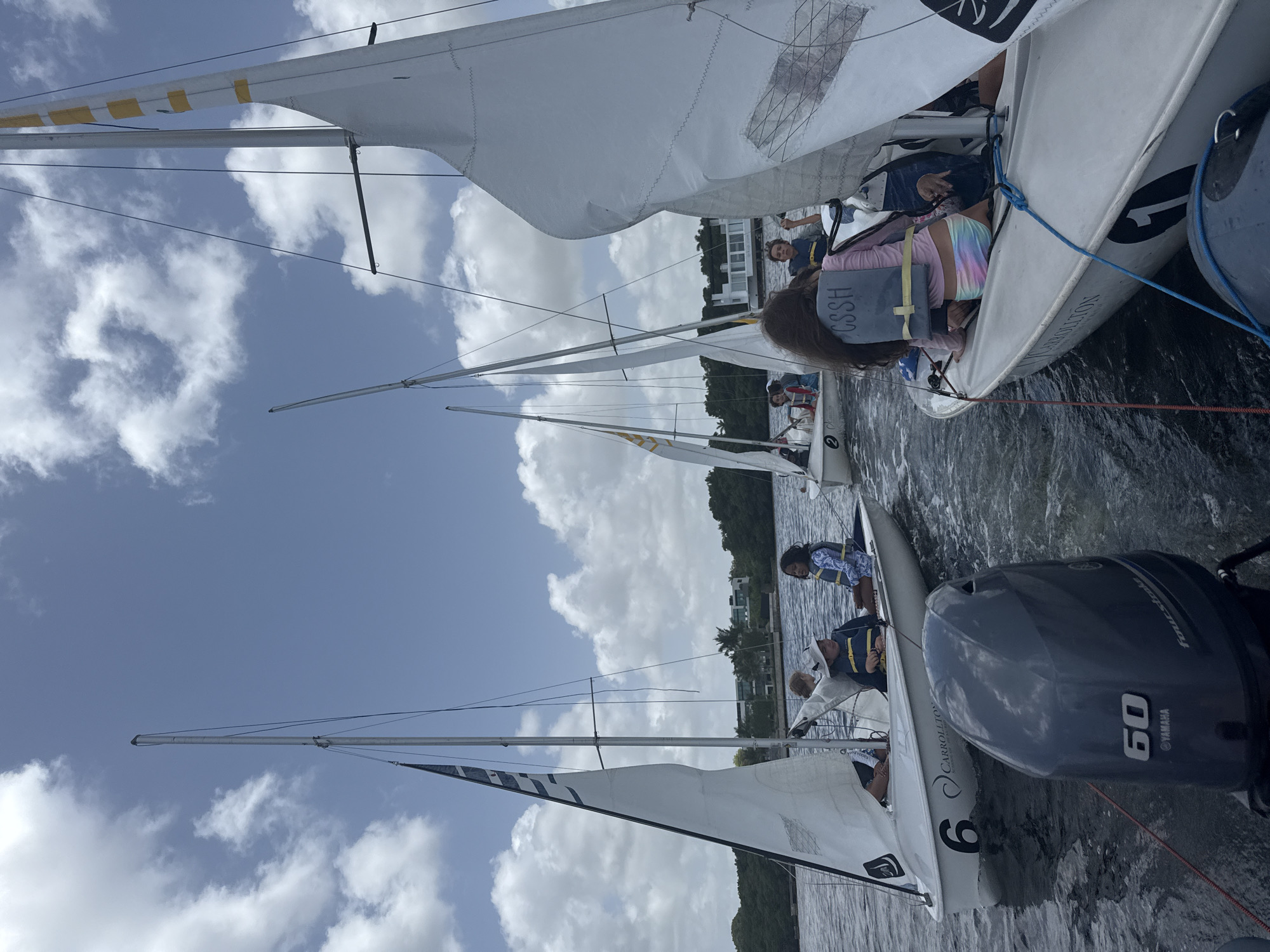 This image depicts a group of children engaged in a sailing activity on a body of water, likely a bay or canal, near Miami, Florida, USA. Several small sailboats, each with at least one child aboard, are visible. One sailboat in the foreground, numbered "6" and bearing the text "CARROLLTON," has an outboard motor attached to its stern. Another sailboat, numbered "1," is also in the foreground and features a child wearing a life vest with "CSSH" printed on it. The children appear to be participating in a sailing lesson or recreational activity. The environment includes calm water, a shoreline with lush vegetation and modern architecture in the background, and a bright, cloudy sky. The overall scene suggests a sunny day conducive to water sports.