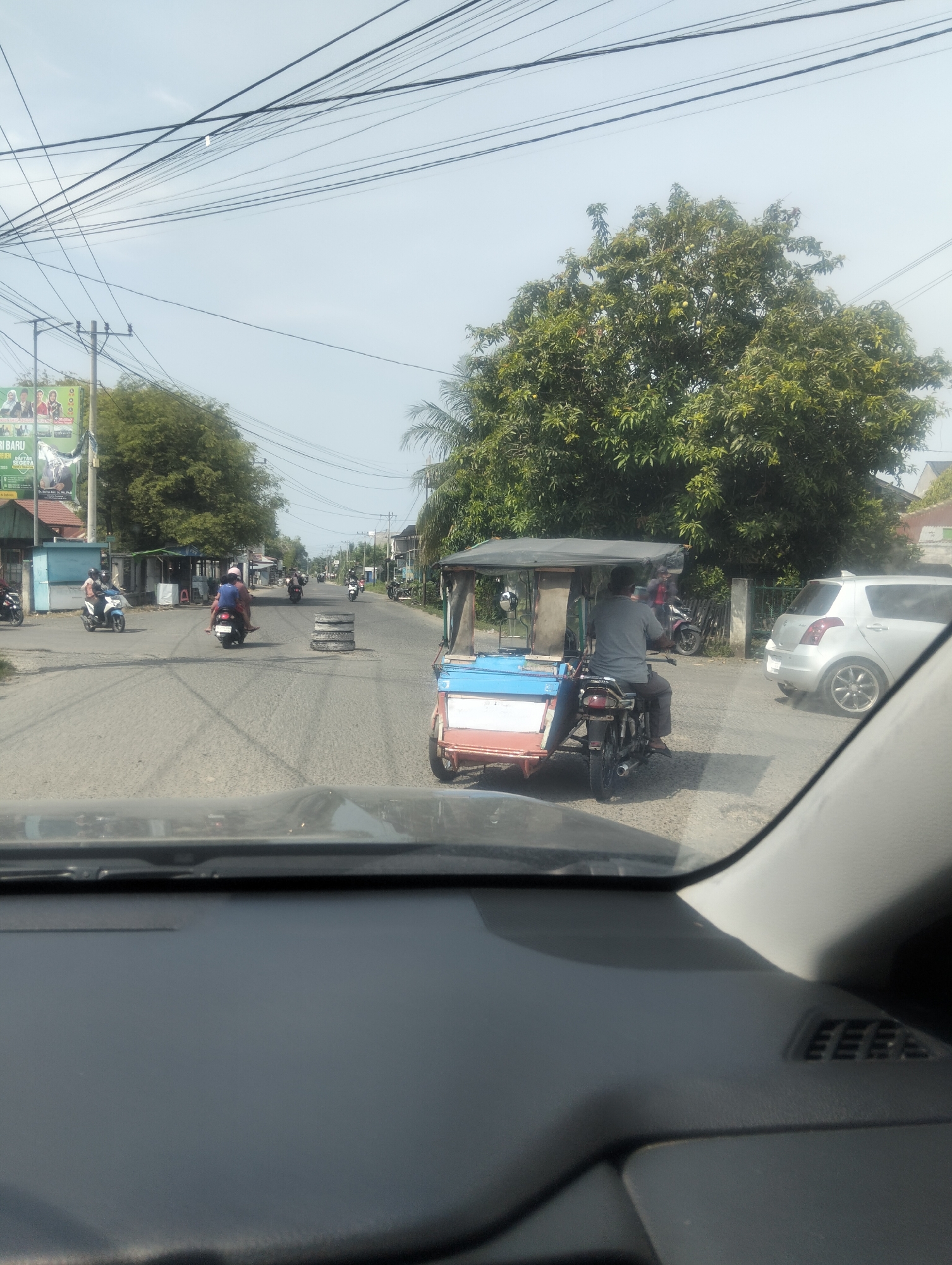In Geulanggang Teungoh, Indonesia, this image captures a street scene with multiple vehicles and pedestrians. A tricycle, also known as a bemo, is prominent in the foreground, with a driver and passenger. Several motorcycles are also on the road, some carrying multiple people. A white car is parked on the side of the street, and there are various buildings and trees lining the road. Power lines are visible overhead, crisscrossing the sky. The road appears to be paved, and there are some tire barriers in the middle of the street, possibly indicating a temporary obstruction or construction. The overall impression is one of a busy, typical day in a local Indonesian town.