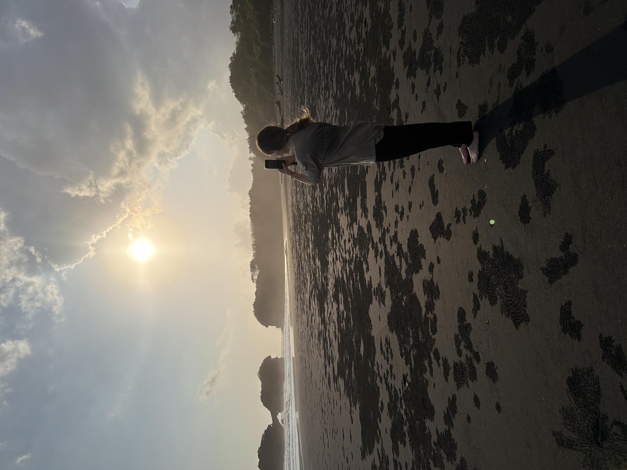 A woman stands on a dark sand beach, facing away from the camera and towards the ocean, holding a smartphone to take a photograph. The sun is setting, casting a warm glow over the scene. In the distance, rock formations rise out of the water, and a lush green hillside borders the beach. The sand is covered in circular patterns, likely made by crabs. The woman is wearing a light-colored t-shirt and dark pants. The sky is filled with soft clouds, some illuminated by the setting sun. The image appears to be taken at sunrise or sunset on a beach in Selemadeg, Indonesia, known for its black sand beaches and dramatic rock formations.