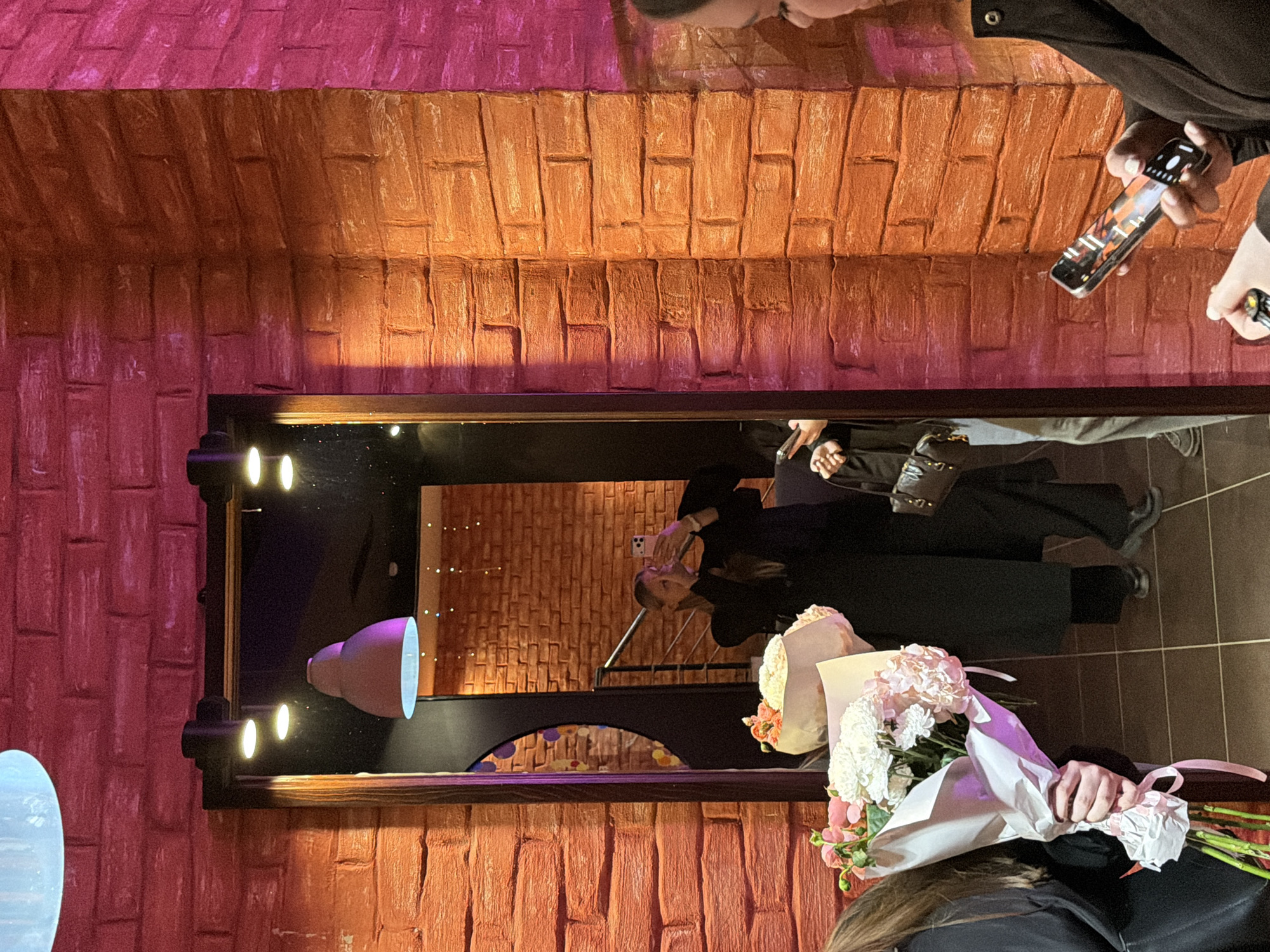 An indoor scene is captured with warm, dramatic lighting, featuring a large mirror and textured walls, likely in Izhevsk, Russia. The image appears to be rotated 90 degrees counter-clockwise.
When correctly oriented, the foreground on the right shows a person, visible from the shoulder up with brown hair, holding a large bouquet of pink and white flowers wrapped in white paper. To their left, dominating the center, is a large rectangular mirror with a dark frame.
The mirror's reflection reveals another person, possibly the same individual, holding a smartphone up as if taking a selfie or recording. This person wears dark attire and has fair hair. Another person is partially visible behind them in the reflection. Within the reflection, bright light fixtures with multiple bulbs and a distinct purple or pink decorative lamp are visible. A smaller, arched opening or mirror is also reflected below these elements.
The walls surrounding the mirror, both to its left and above it, are highly textured, resembling stacked bricks or stones, painted in a palette of reddish-orange, fading into deeper reds and pinks in various sections due to the lighting. Parts of other individuals, mainly hands and arms, are visible in the upper right portion of the image, one holding a smartphone with its screen visible, and another holding a small, unidentifiable object.
The activity suggests a social gathering or event, with people interacting, posing, and taking photos. The soft, varied lighting creates an intimate evening atmosphere. No explicit text is visible in the image.