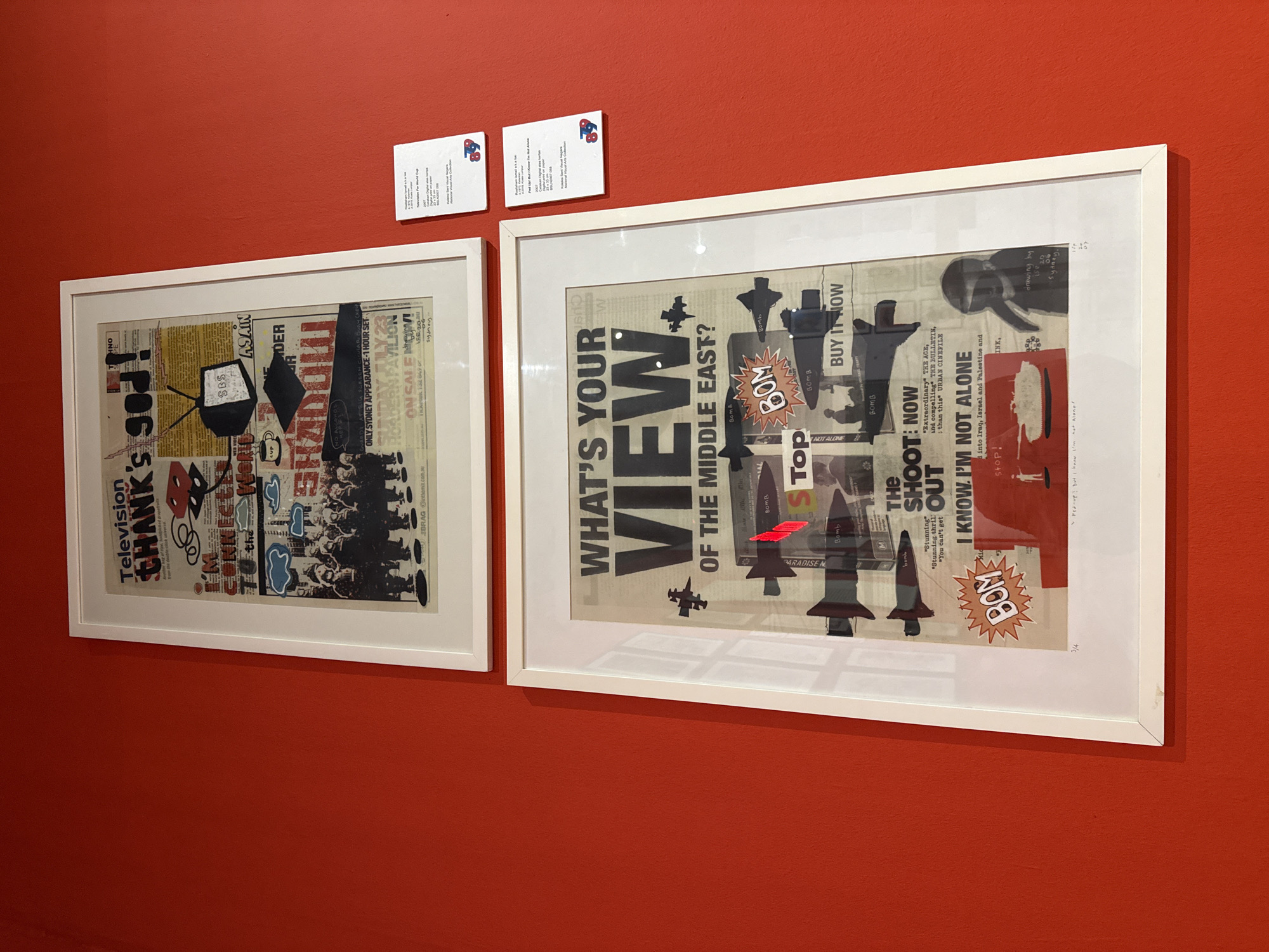 Two framed artworks hang on an orange wall. The artwork on the left contains the text "Television THANK's god! CONNECT to the..." and includes images of a television, coffee cup, and people. The artwork on the right contains the text "WHAT'S YOUR VIEW of the MIDDLE EAST?" and includes images of missiles, airplanes, and explosions. There are two small white information cards above each artwork. The location is not Kuala Lumpur, Malaysia.
