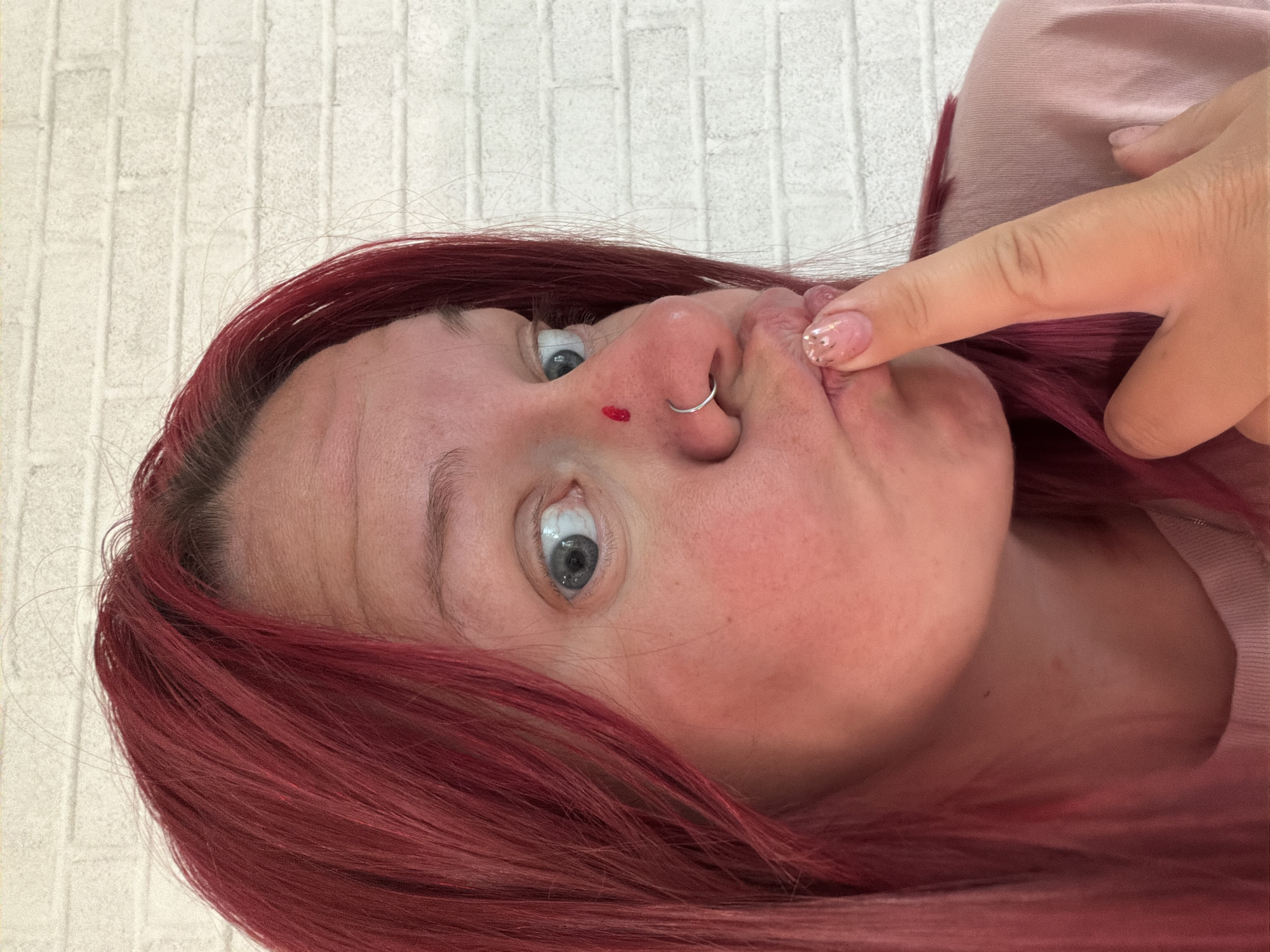 A selfie angled upward towards a woman with red hair. The background is a white brick wall. The woman has blue eyes, a nose ring, and a small amount of blood on her nose. Her index finger is touching her upper lip. She is wearing a light pink shirt. The location in the city of Chaikovsky, Russia cannot be determined from the image.