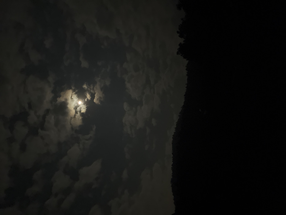 The scene depicts a nighttime landscape in La Marquesa, Mexico. The sky occupies the upper two-thirds of the image, presenting numerous scattered cumulus clouds illuminated by a bright, full moon located slightly right of the upper center. The moonlight creates varying shades of gray within the clouds, contrasting with the dark portions of the sky. The lower third of the image consists of a completely dark, undulating silhouette of a forested hillside or mountain range, devoid of specific detail due to the lack of light. The immediate foreground is entirely obscured by darkness. No people, man-made objects, actions, or interactions are discernible.