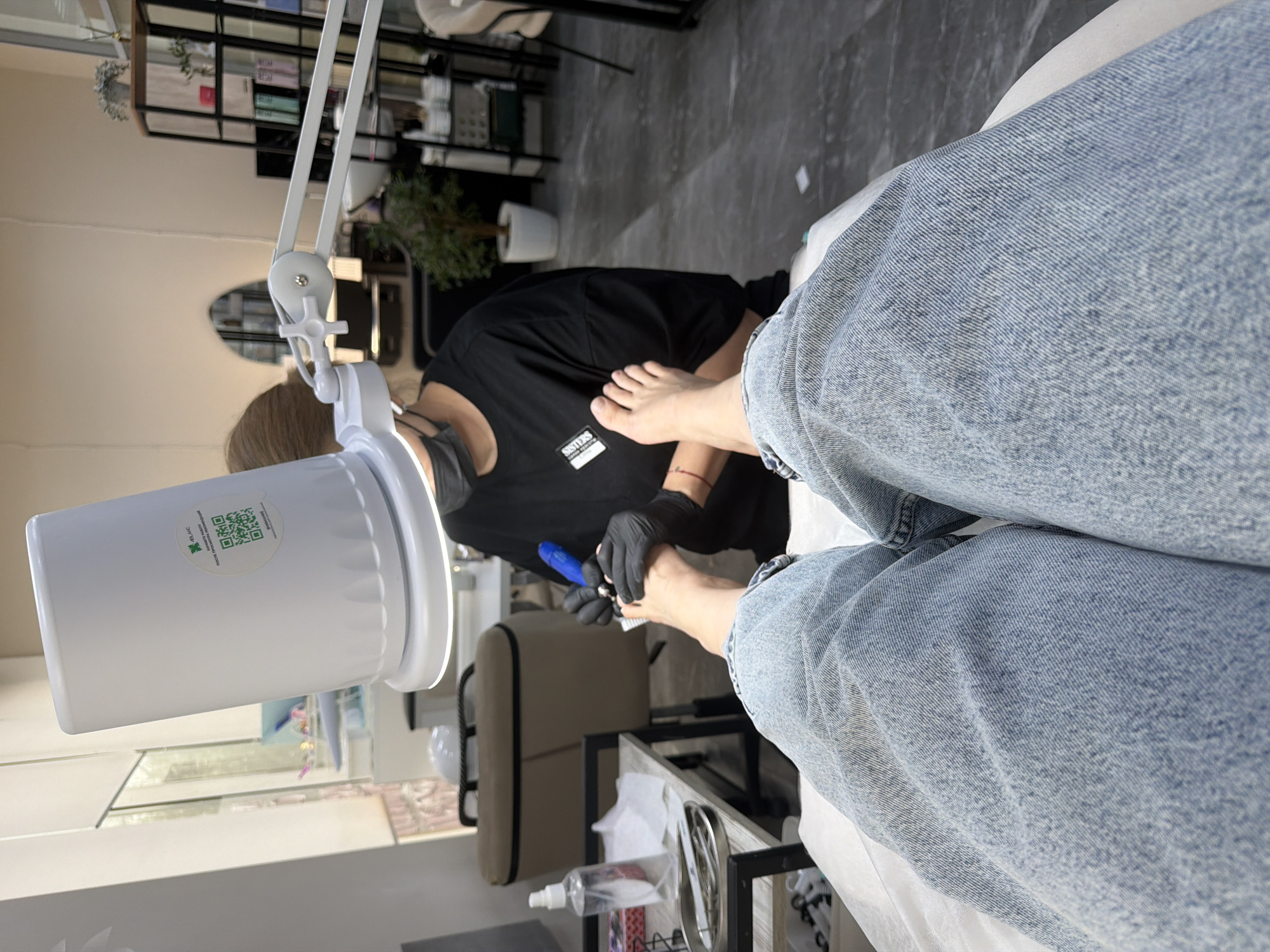Here is a detailed description of the image:
**Scene Description:** The image captures a scene of a person receiving a pedicure, likely indoors in a salon. The camera angle is positioned from the point of view of the client, looking down towards the pedicurist and their own leg.
**People:**
* There is a person getting a pedicure, wearing blue jeans. The person's leg and foot are prominently visible.
* There is a manicurist or pedicurist performing the service. They are wearing black gloves and a black shirt and a face mask.
**Objects:**
* A white lamp with an attached white cylindrical object, potentially a light or device for the pedicure.
* Pedicure tools (a blue tool is visible)
* Shelves and furniture in the background.
* A bottle of liquid disinfectant is on a shelf.
**Actions/Interactions:**
* The pedicurist is holding and working on the client's foot.
* The person is sitting or reclining while receiving the pedicure.
**Environment:**
* The background includes furniture and equipment typically found in a salon, like shelves.
* The location appears to be well-lit and clean.
* There is no information in this picture to confirm that this picture was taken in Moscow, Russia.