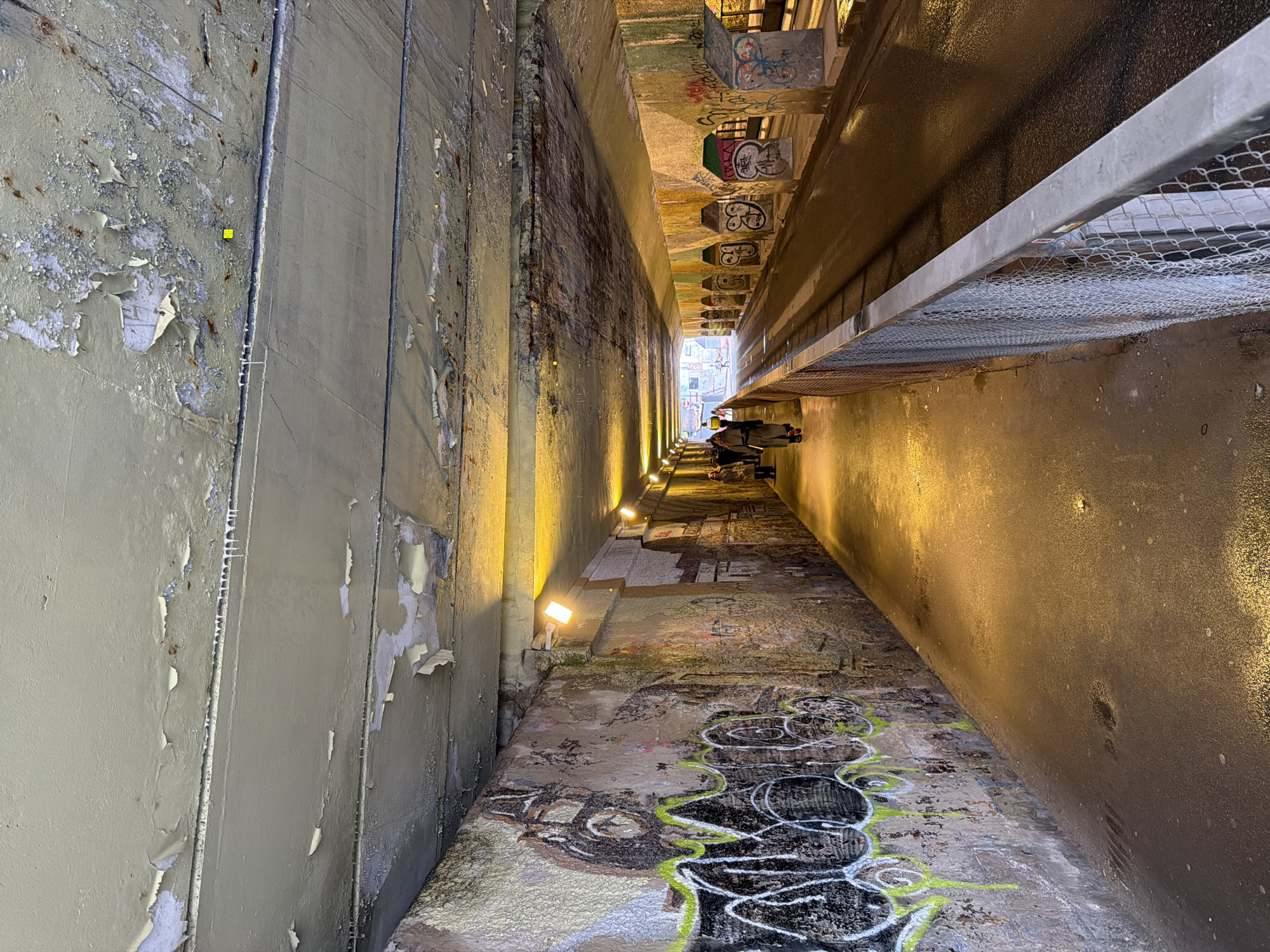The image captures a gritty urban underpass or tunnel, viewed from one end looking towards its upward-sloping exit. The concrete walls and overhead structures define a narrow, elongated pathway.
The left wall shows significant wear, with large sections of light greyish-green paint peeling away, exposing darker layers and some rust streaks beneath. On the right side, above the pathway, an overhead structure is supported by several rectangular concrete beams, all heavily adorned with various graffiti tags and abstract markings. Below this structure, a metal railing with a mesh or chicken wire fence runs along the upper part of the wall.
The concrete pathway itself is extensively covered in layers of diverse graffiti, featuring swirling patterns, outlines, and abstract designs in white, black, grey, and vibrant neon green. Several light fixtures are strategically mounted low on the walls, casting a warm, yellow-orange glow that illuminates the path and parts of the walls, creating a striking contrast with the raw concrete and graffiti.
Towards the far end of the underpass, where the path inclines towards the exit, several individuals are visible. They appear to be dressed in coats, suggesting cooler weather. The tunnel opens up to a brighter outdoor scene with buildings in the distance, indicating an exit onto a street or urban area.
The artificial lighting within the underpass suggests the photograph was taken during the evening or night. The overall atmosphere is utilitarian and urban, yet softened by the warm lighting and vibrant, albeit unpolished, street art. While numerous graffiti tags are present, no specific legible text is clearly discernible. The scene is located in Moscow, Russia.