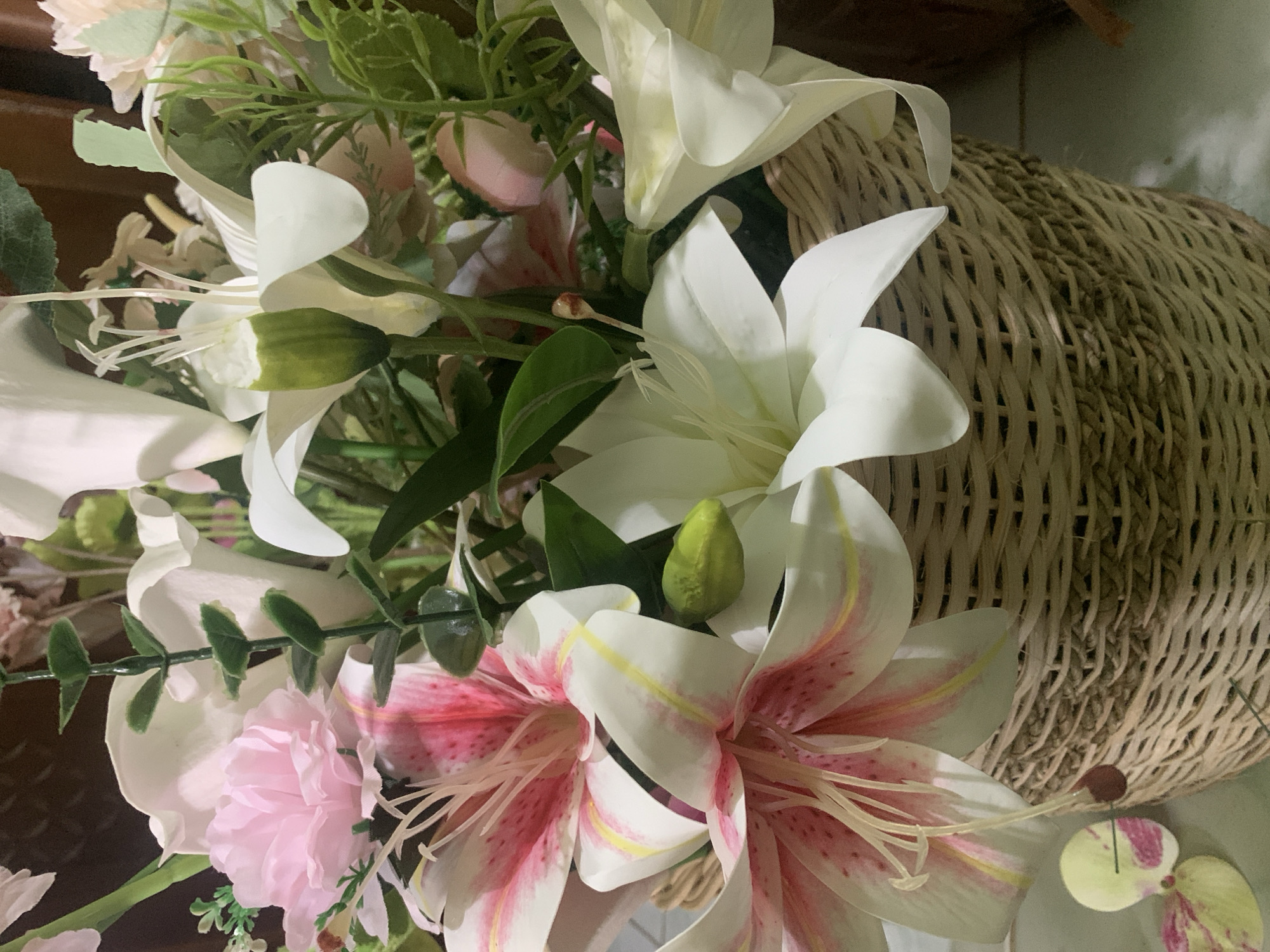 The image shows a close-up of a woven basket filled with artificial flowers. The flowers are a mix of white lilies with pink and yellow accents, and soft pink carnations. Green artificial foliage is also present, adding to the fullness of the arrangement. The basket is intricately woven with a light brown, natural material. The background is blurred, but appears to be indoors with a wooden surface visible on the right. The lighting suggests a natural light source from the left, casting soft shadows. The scene is static and does not depict any people or actions. The location in Kuala Pembuang, Indonesia, is not visually discernible from the provided image.