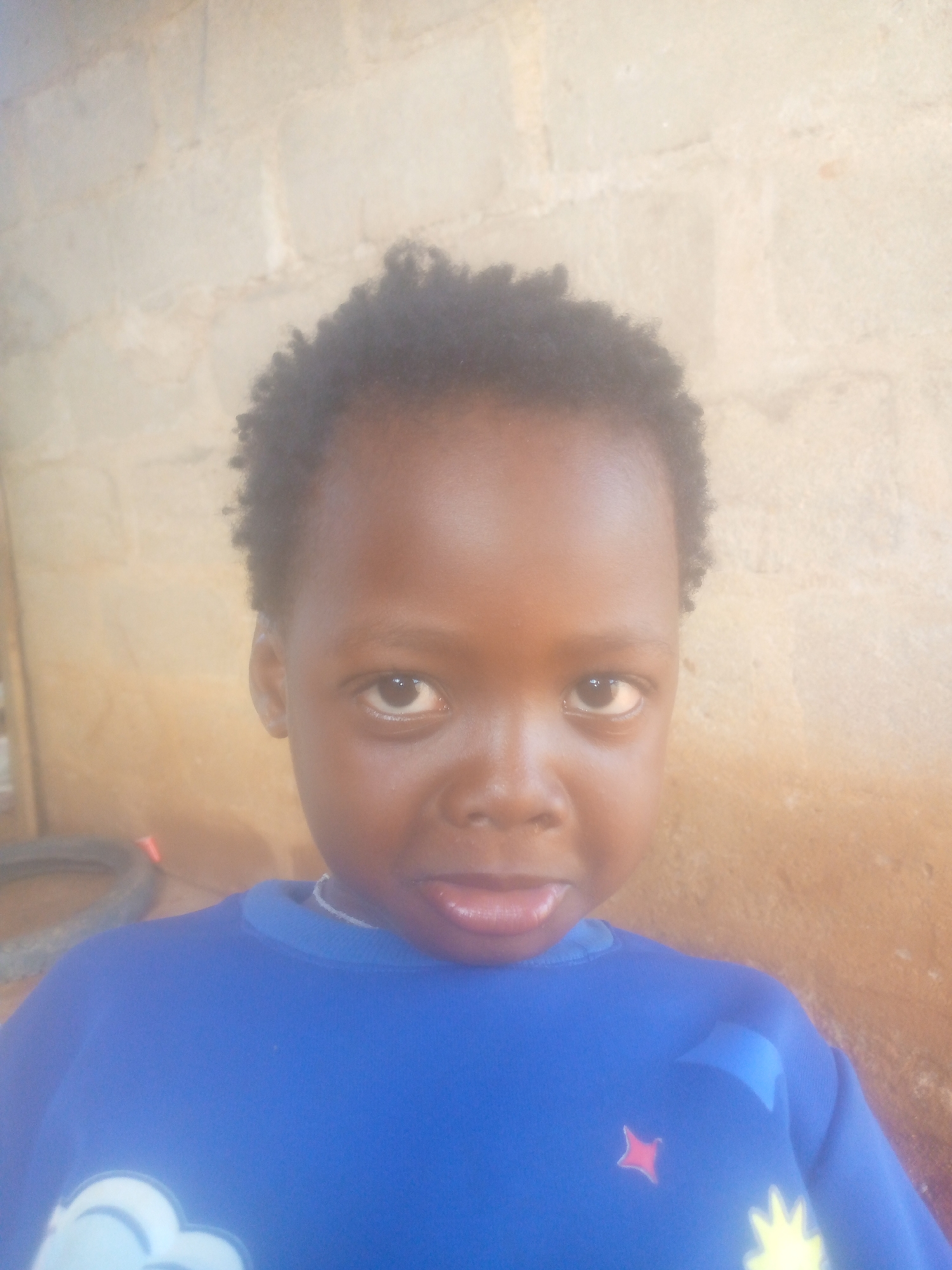A close-up portrait shows a young child with dark skin, dark curly hair, and dark eyes looking directly at the camera. The child's mouth is slightly open, with the tongue visible. They are wearing a long-sleeved blue shirt adorned with light-colored designs, including what appears to be a white cloud shape on the lower left and a yellow star-like shape and a small red star on the right side of the chest. A thin, light-colored necklace is visible around the child's neck.
The background consists of a wall made of light-colored, possibly painted, bricks or concrete blocks, giving a somewhat textured appearance. On the far left side of the image, partially visible behind the child's shoulder, is a dark, circular object that might be a tire or a coiled hose, and a small hint of red color. The lighting suggests it is daytime, with natural light illuminating the scene. The child's expression is neutral, engaging the viewer directly. No text is visible in the image.