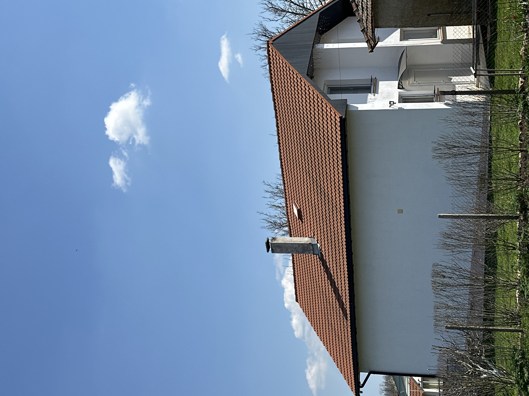 The image shows a two-story house in Balkantsi, Bulgaria. The house is white with a red-tiled roof and a chimney. A narrow fence runs along the side of the house, separating it from a grassy area with small bushes. The sky is blue with a few scattered clouds. There are no people visible in the image.