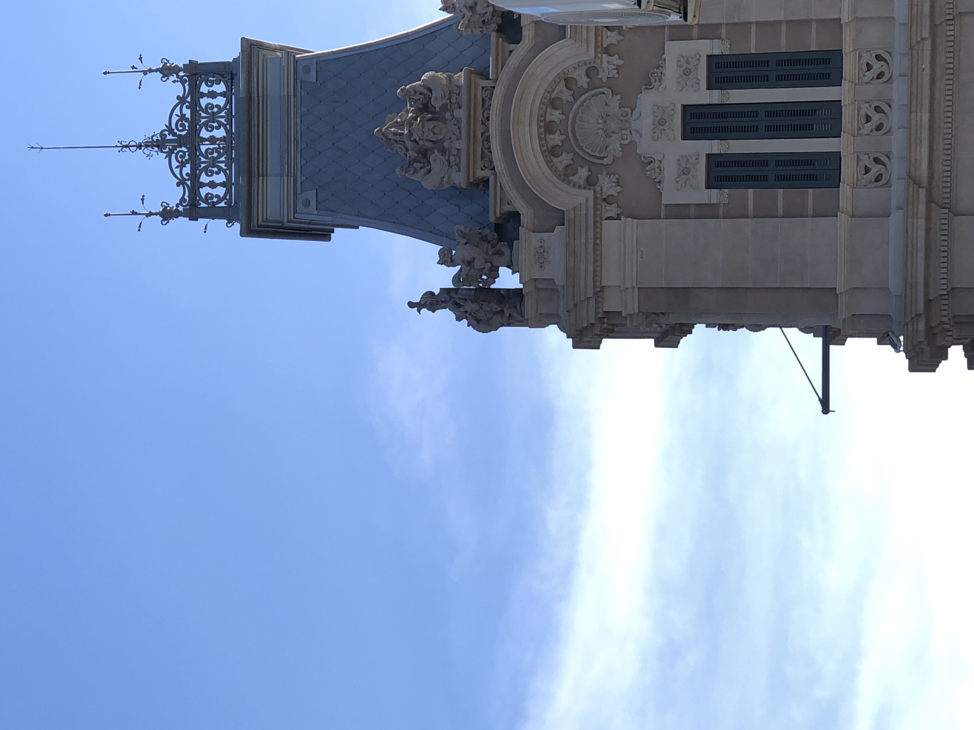A low-angle shot captures the upper portion of a stone building with a grey, mansard-style roof against a bright blue sky with wispy clouds. The building's facade is adorned with ornate architectural details, including sculpted figures, decorative moldings, and arched window frames. Three dark green shutters are visible on a prominent window. The building's spire is capped with an intricate metalwork railing and a lightning rod.
