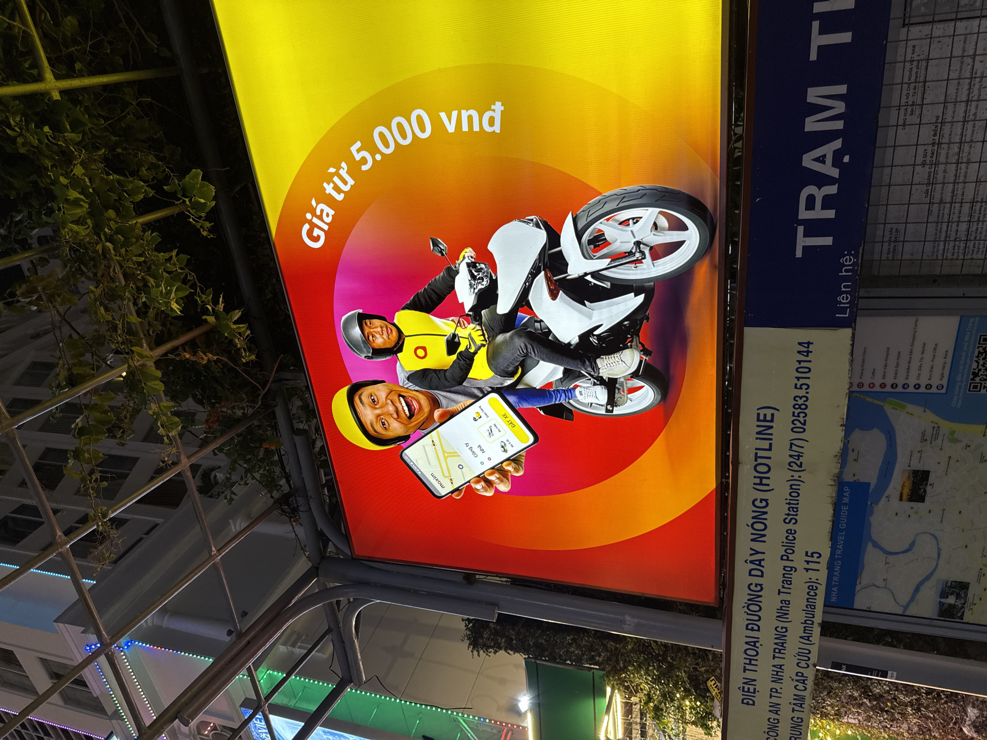 An illuminated billboard at night in Nha Trang, Vietnam, features two men on a white motorcycle against a vibrant red and yellow background. The man in front, wearing a yellow helmet and jacket, holds up a smartphone displaying a navigation app with Vietnamese text. The man behind him, wearing a black helmet and yellow jacket, looks ahead. The billboard advertises a service with text in Vietnamese, including "Giá từ 5.000 vnd," which translates to "Price from 5,000 VND." Below the billboard, a blue sign provides contact information, including a "HOTLINE" for the Nha Trang Police Station and Ambulance, along with a section titled "NHA TRANG TRAVEL GUIDE MAP" and another sign with Vietnamese text that partially reads "TRẠM TH". The scene is set against the backdrop of a building with lights and some greenery.