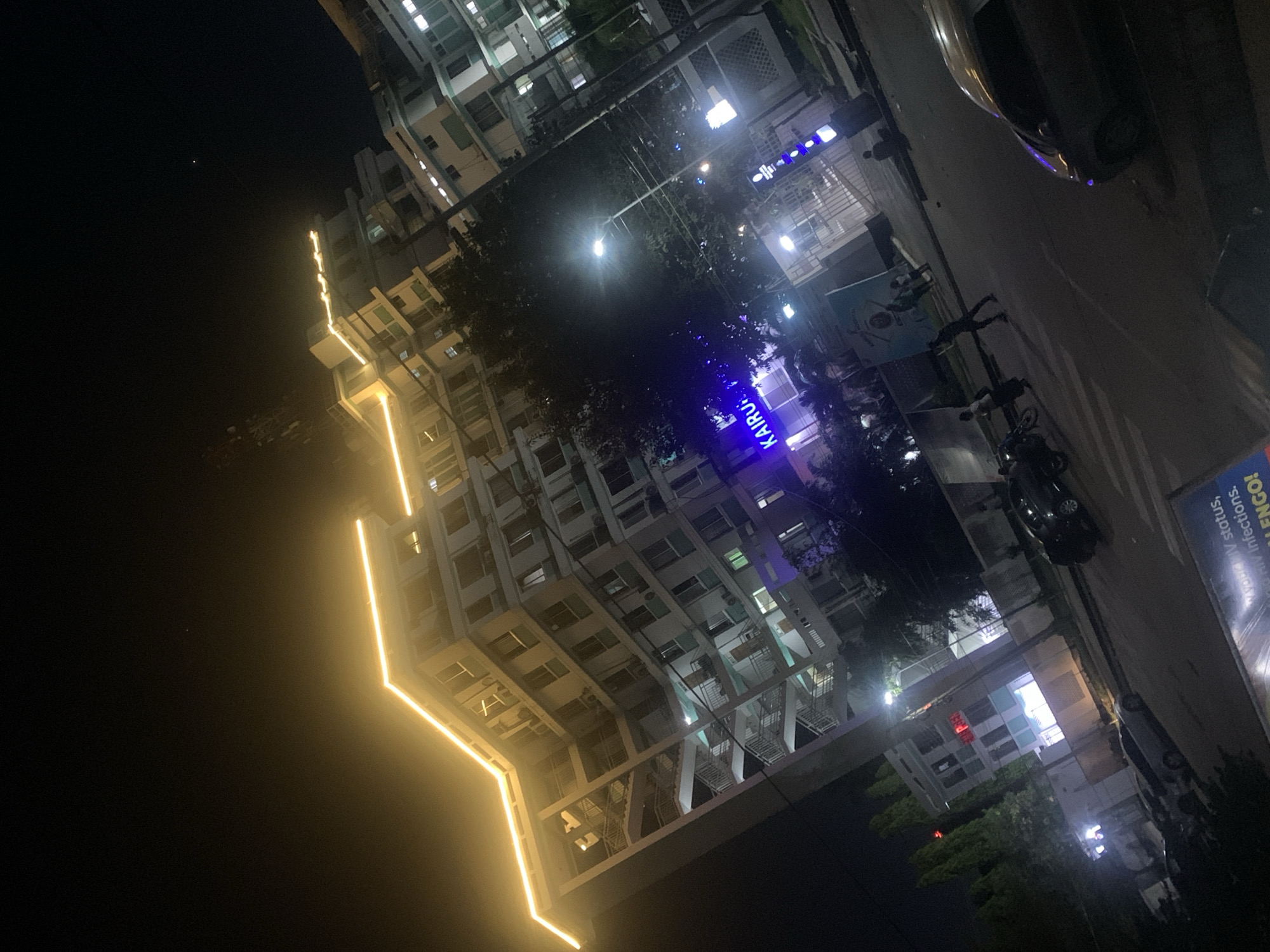 At night, the city of Dar es-Salaam, Tanzania, is depicted at street level, rotated slightly clockwise. A tall building with a jagged yellow-lit edge is illuminated. The windows, mostly lit, reveal floors. Streetlights provide general illumination, and a sign reads "KAIRU" in purple neon. Trees fill the space between buildings, and a car is parked to the right. Several motorcycles are also visible. A sign promoting HIV testing can be seen. The sky appears dark.