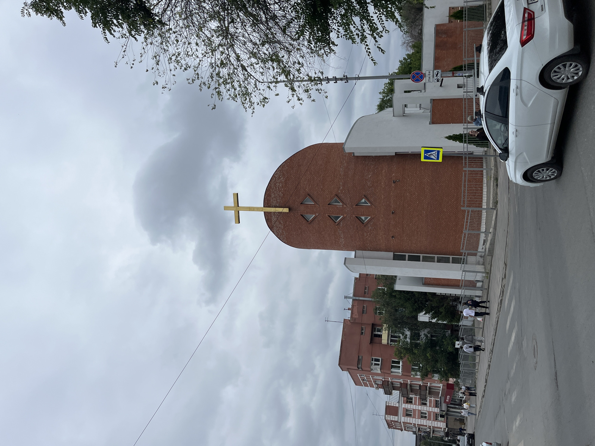 The image shows a street scene in Saratov, Russia, with a modern church featuring a gold cross prominently on its brick facade. The church is adjacent to several residential buildings and a paved street with a crosswalk. A white car is visible in the foreground, and several pedestrians are walking on the sidewalk. The sky is overcast, suggesting a cloudy day. The overall impression is a typical urban environment in Russia.