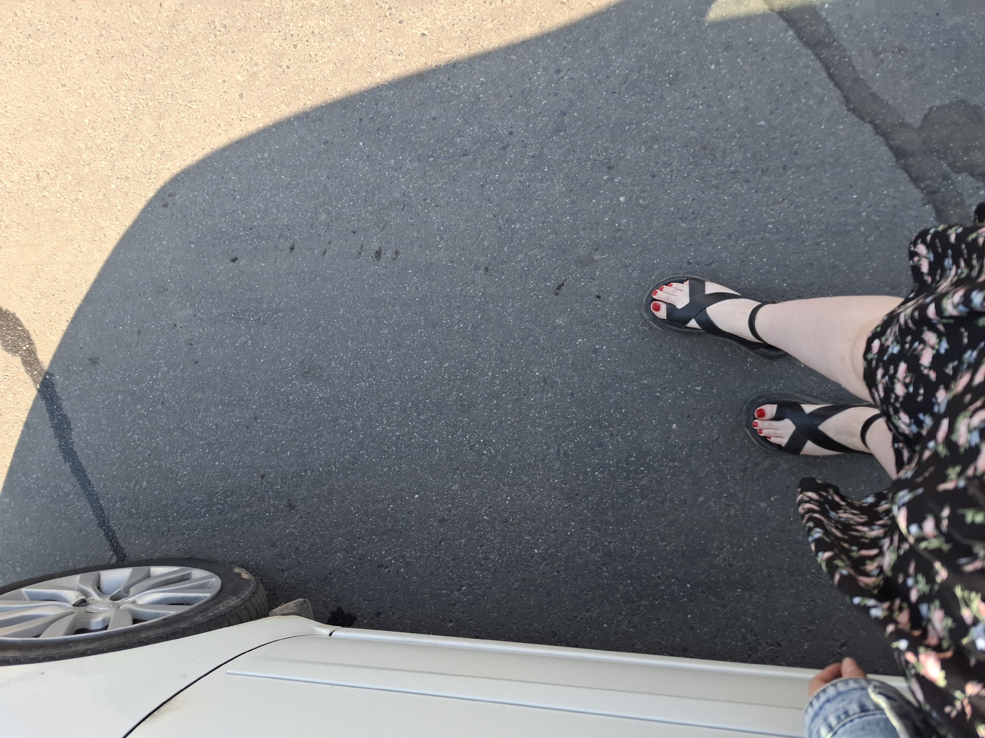 The image shows a person standing on asphalt next to a white car. The person is wearing black sandals and has red nail polish on their toes. They are also wearing a floral dress and a denim jacket. The car's front wheel is visible in the bottom left corner of the image. The asphalt surface is partly in sunlight and partly in shadow. The location is not identifiable as Izhevsk, Russia, from the visible context.