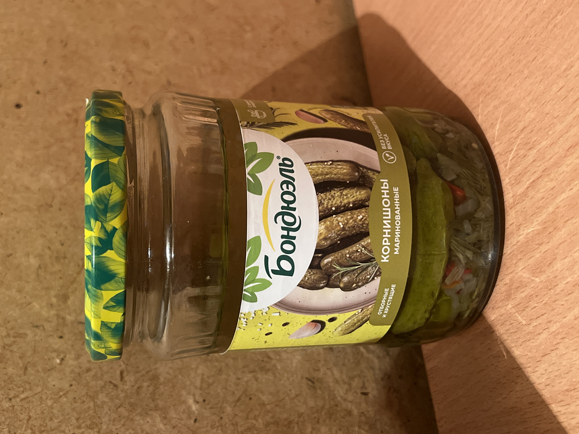 A glass jar of "Bondjuel" brand cornichons sits on a wooden surface. The jar is filled with pickled cucumbers and brine, and features a yellow label with green leaf patterns. The label displays the brand name in large, white letters, along with an image of cornichons on a plate, and text in Russian that translates to "Cornichons, marinated" and "Selected and crunchy." A green and yellow patterned lid seals the jar. The background is a textured, light brown surface. The lighting suggests an indoor setting, with a shadow cast on the wooden surface by the jar. There is no indication of the city of Iksha, Russia, or any people present.