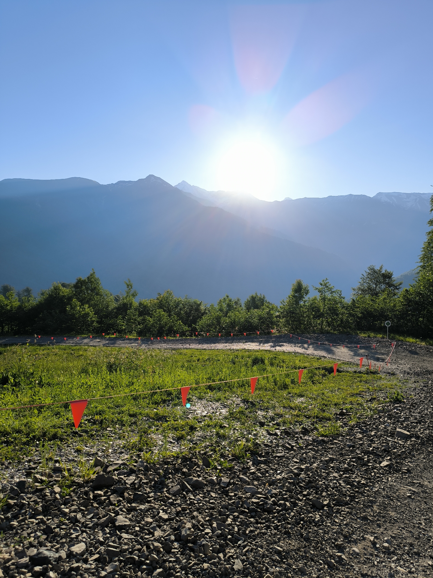 A bright, sunlit image captures a mountain landscape in Estosadok, Russia. The sun is positioned high in a clear blue sky, with rays of light emanating outward, creating a lens flare effect. In the foreground, a gravel path curves to the right, bordered by lush green grass. A yellow rope stretches across the scene, marked by a series of small, red triangular flags. Behind the grass, a line of dense green trees forms a backdrop. In the distance, majestic mountains rise, their peaks silhouetted against the bright sky, with some appearing to have snow cover. The overall scene suggests an outdoor recreational area, possibly a hiking or biking trail, set within a natural mountainous environment.